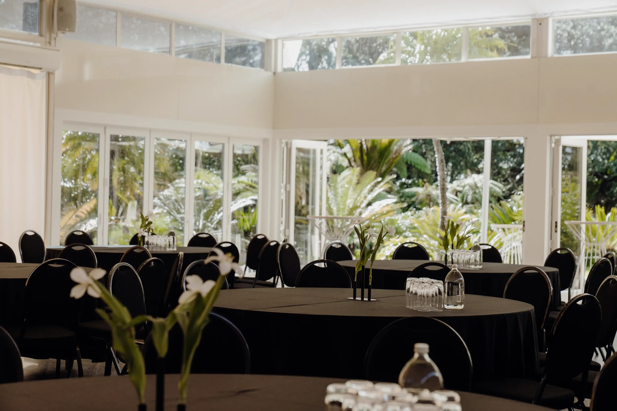 An indoor banquet hall with round tables covered in black tablecloths, set with glassware, and decorated with white flowers in vases. Large windows let in natural light and reveal tropical greenery outside.