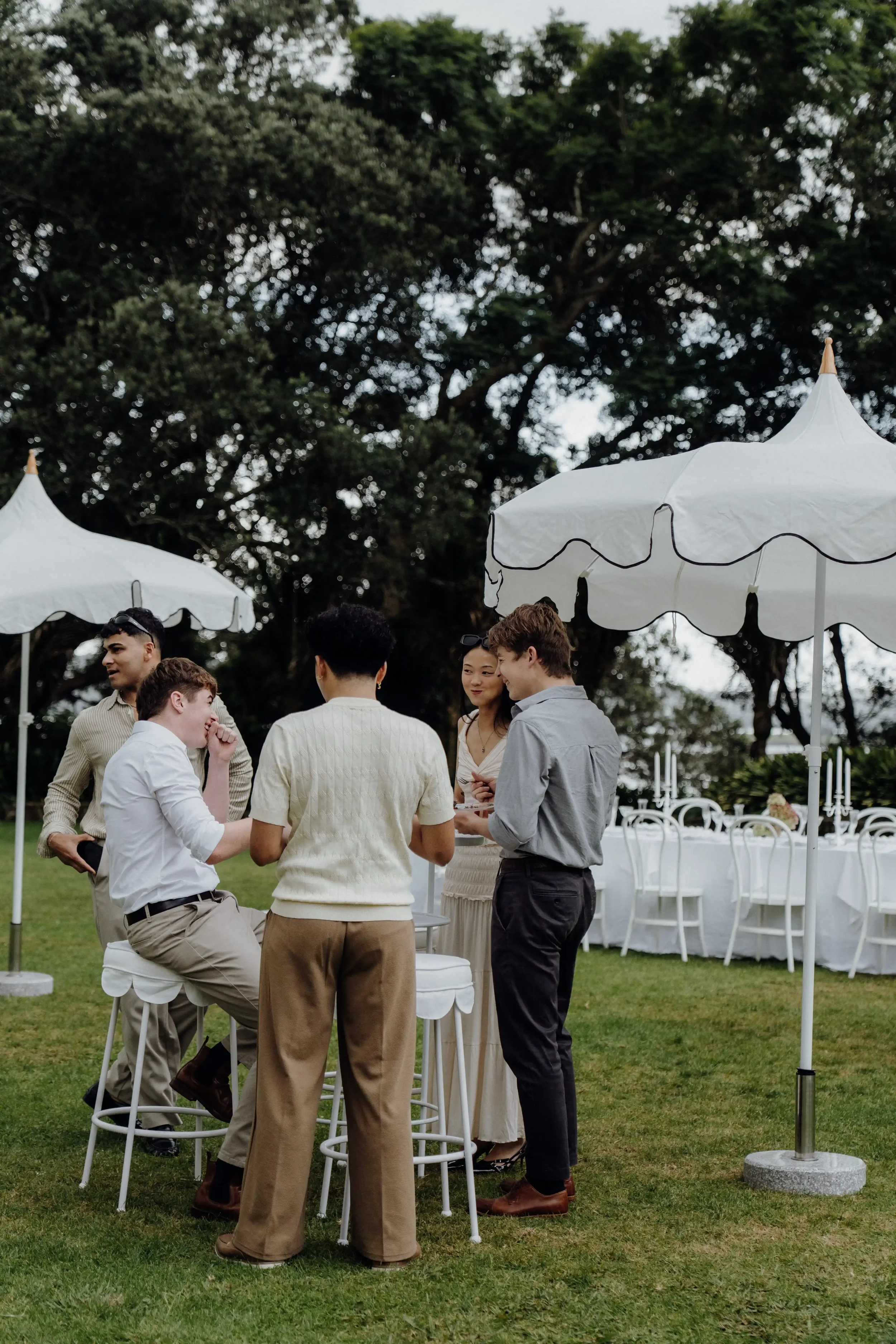 A group of six people, four men and two women, gathered outdoors under a white canopy tent with white chairs and tables, engaged in conversation. The setting is a garden or park with large trees and green grass.