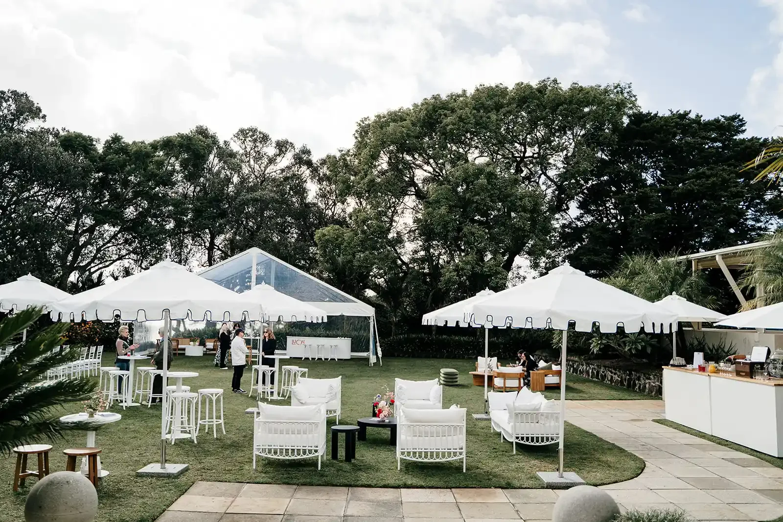 Outdoor event setup with white tents, umbrellas, and seating areas on a grass lawn, surrounded by trees.