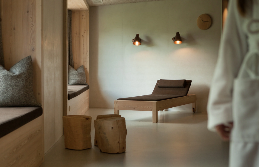 Ein ruhiger Raum mit Holzbetten und Sitzgelegenheiten, moderner Minimalstil, Wandlampen und einer Person in weißem Arztkittel im rechten Bildbereich.