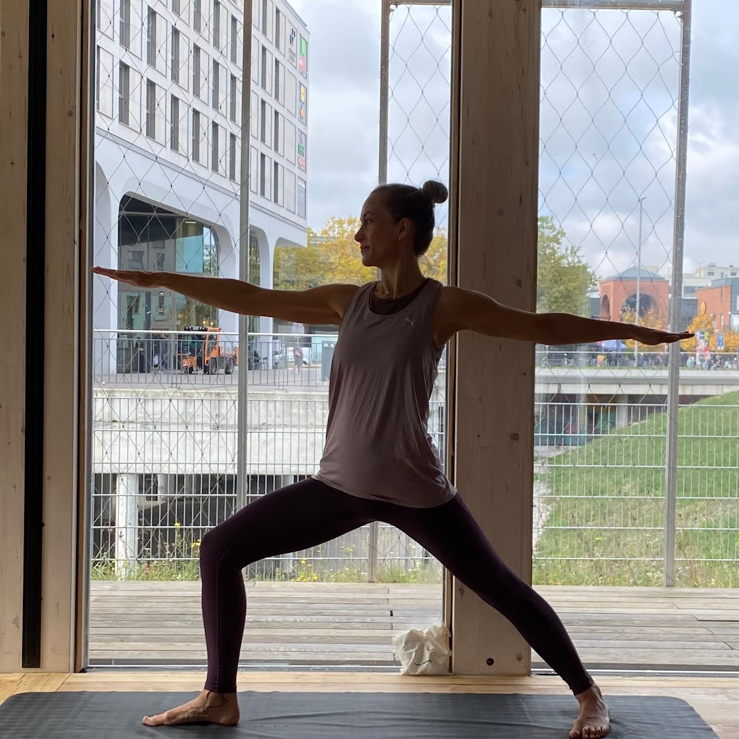 Frau in Yoga-Position mit ausgestreckten Armen und in eine Lordose gebeugtem Bein im Yoga-Studio mit großen Fenstern