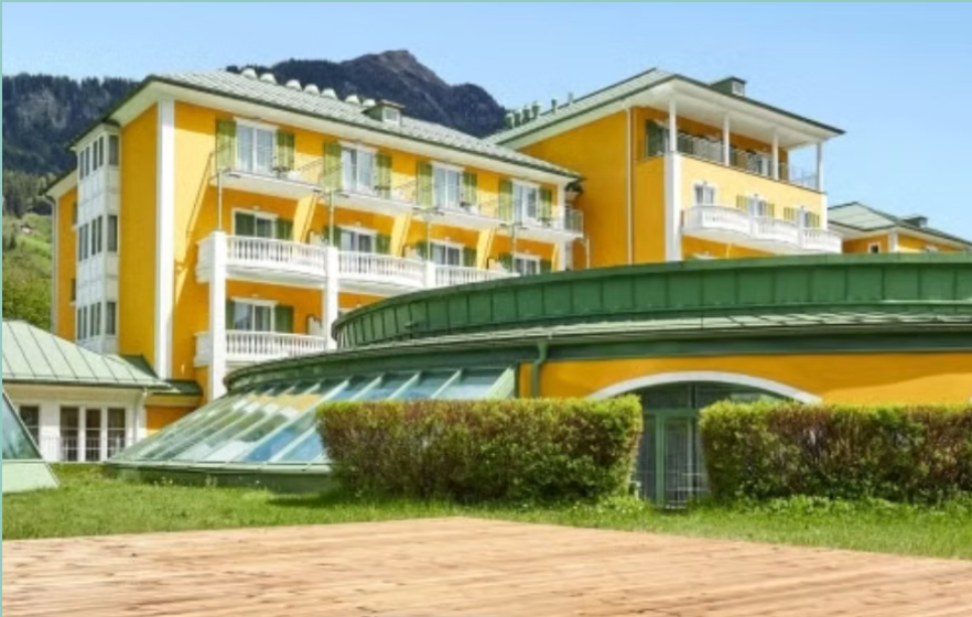 Ein gelbes mehrstöckiges Hotel mit Balkonen, grünem Dach und großen Fensterflächen vor einer bergigen Landschaft.
