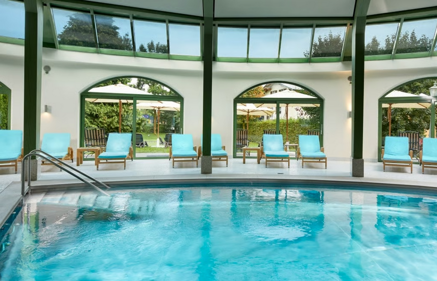 Innenaufnahme eines Schwimmbads mit Liegestühlen und großen Fenstern