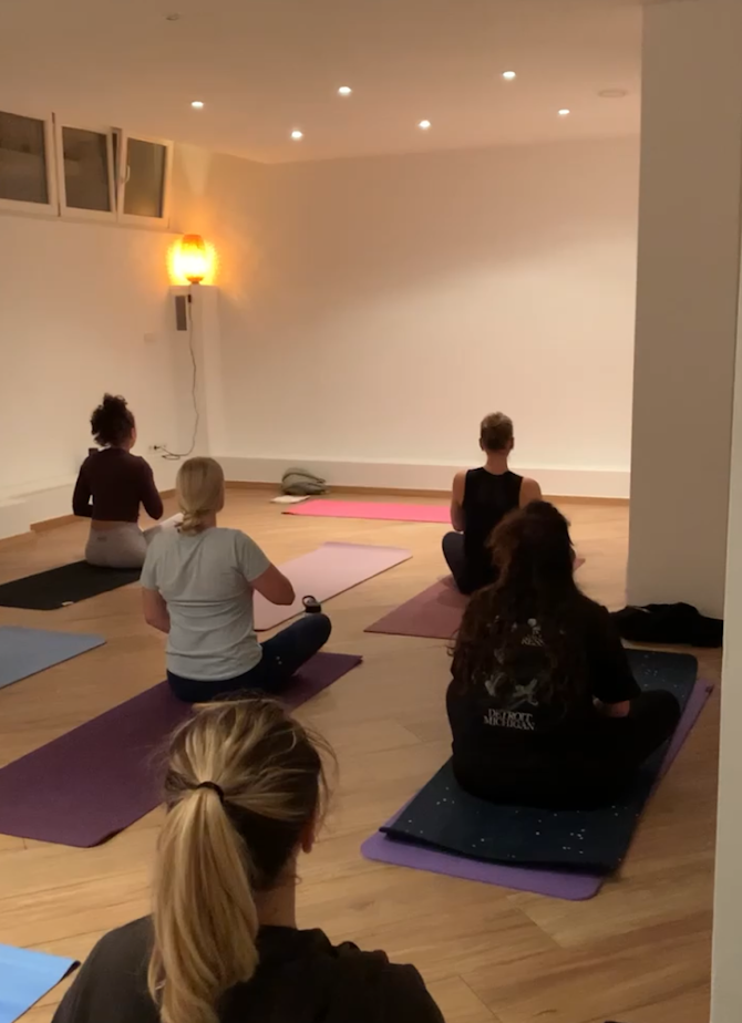 Menschen nehmen an einem Yoga- oder Meditationskurs in einem hell erleuchteten Raum teil, sitzen auf Yogamatten in entspannter Haltung.