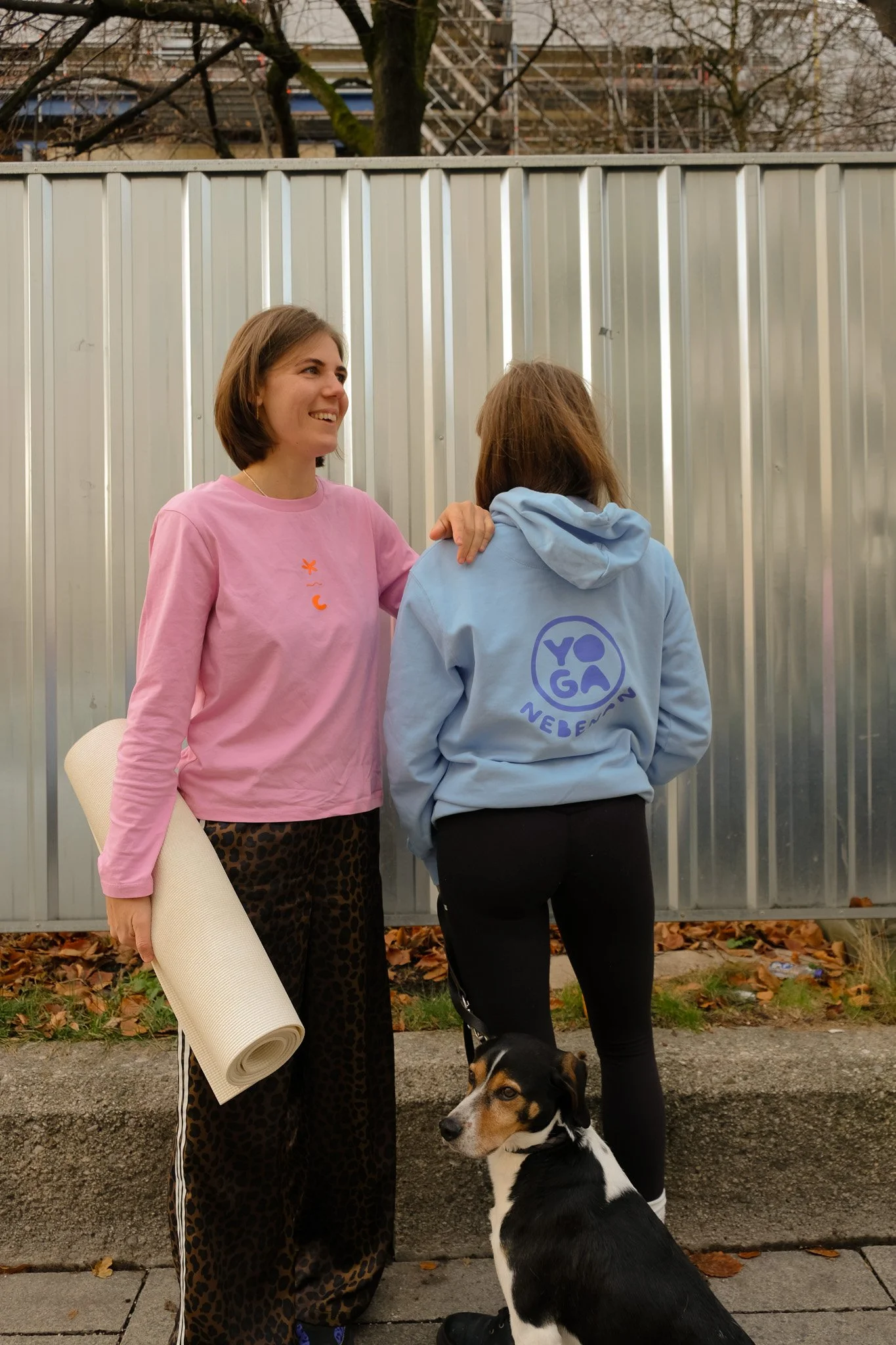 Zwei Frauen stehen einander gegenüber, eine lächelt, während die andere mit dem Rücken zur Kamera steht. Sie sind im Freien, vor einer Metallzaun und fallen in Herbstfarben. Eine der Frauen hält eine Yoga-Megamatte, und ein Hund sitzt an ihrer Seite.