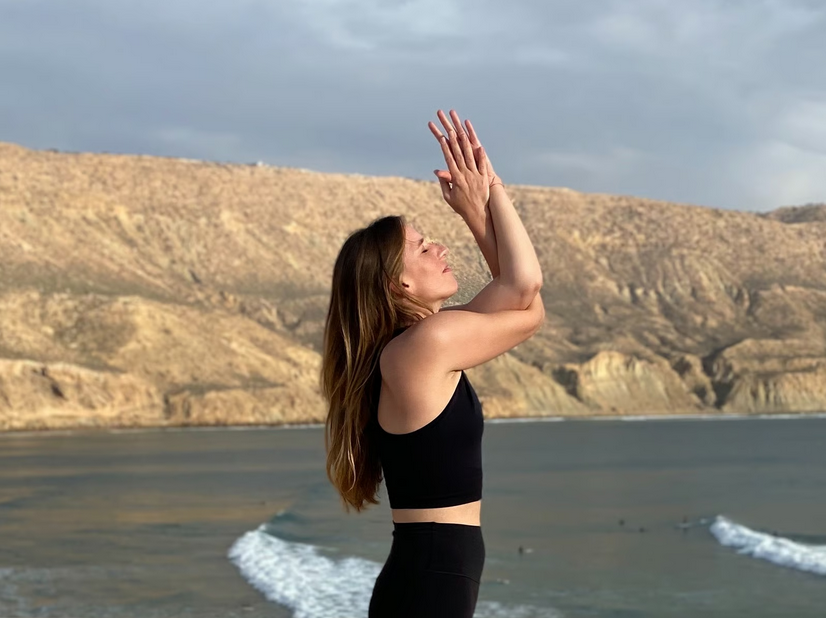 Frau macht Yoga am Strand bei Sonnenuntergang, im Hintergrund Hügel und Wasser.