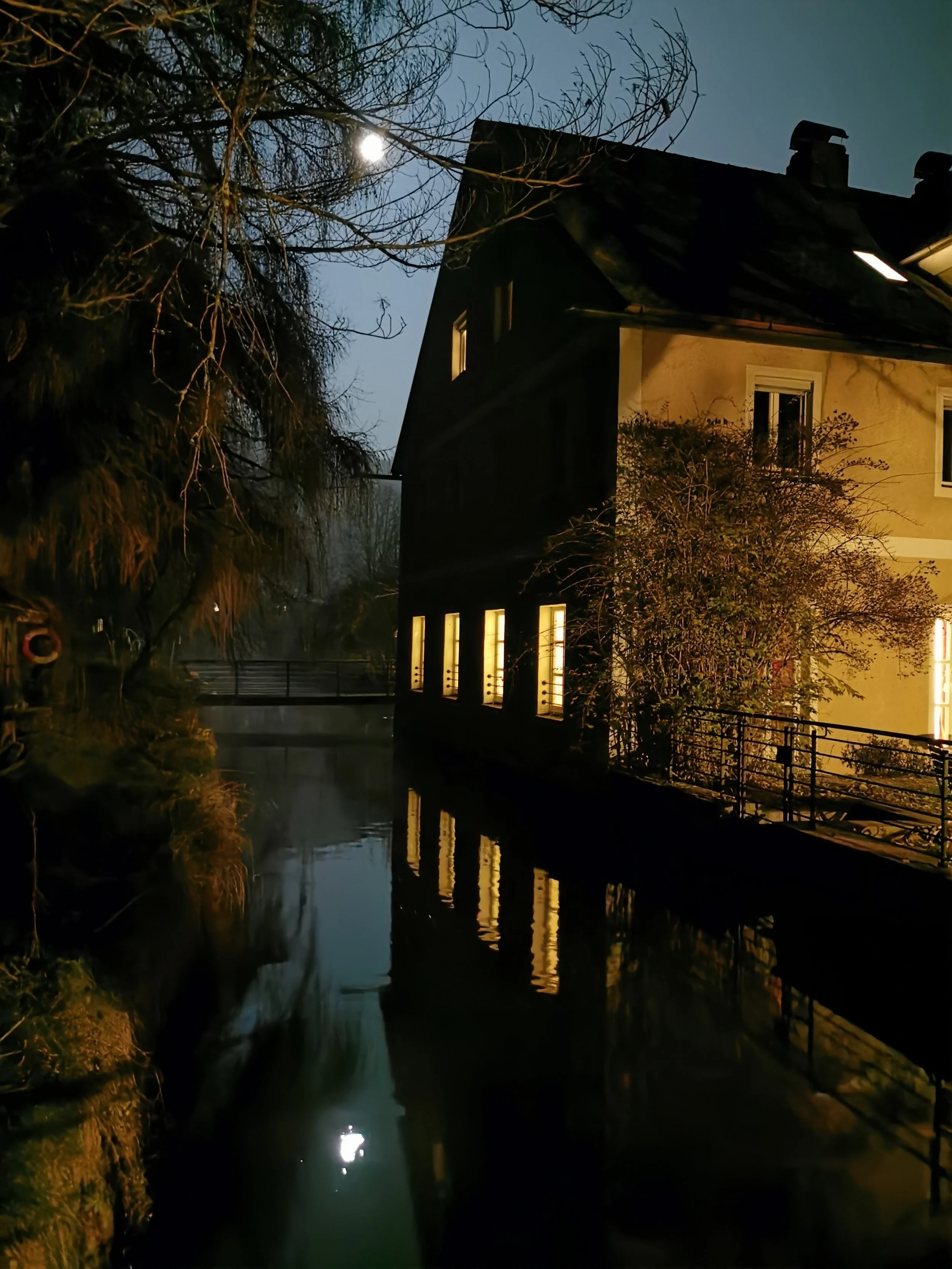 Ein Haus mit beleuchteten Fenstern an einem Fluss bei Nacht, mit kahlen Bäumen im Vordergrund und Mond im Himmel.