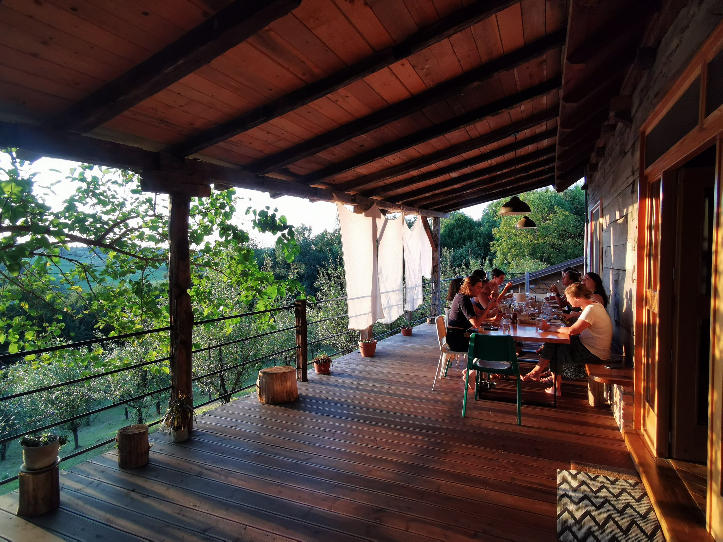 Menschen sitzen an einem langen Holztisch auf einer Veranda mit Wald im Hintergrund, Sonnenlicht fällt durch das Dach. Es gibt Pflanzen und Töpfe auf dem Boden, und die Veranda ist teilweise geschlossen mit einer Steinwand und Fenstern.
