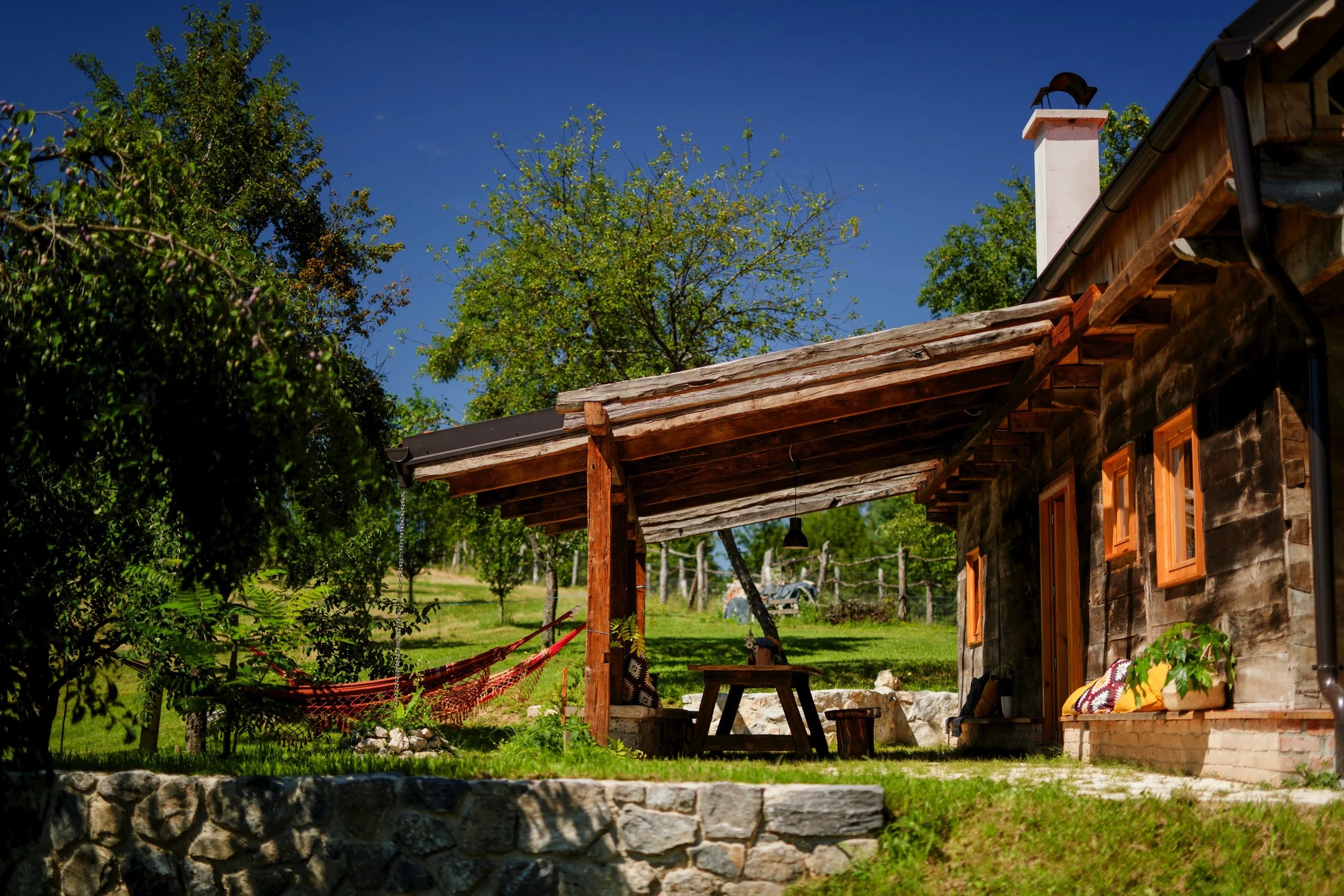 Ein rustikales Haus mit Holzverkleidung, grünen Bäumen und einem blauen Himmel im Hintergrund. Es gibt eine überdachte Veranda mit Holzmöbeln und bunten Kissen, sowie eine Steinmauer im Vordergrund.