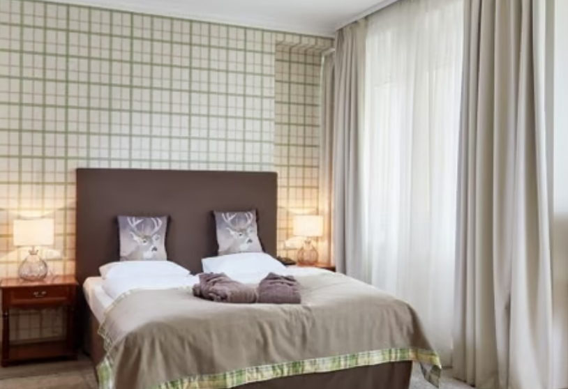 Ein gemütliches Schlafzimmer mit zwei Kopfkissen, einem beige Bett, Nachttischen mit Lampen, und einem Fenster mit weißen Vorhängen.