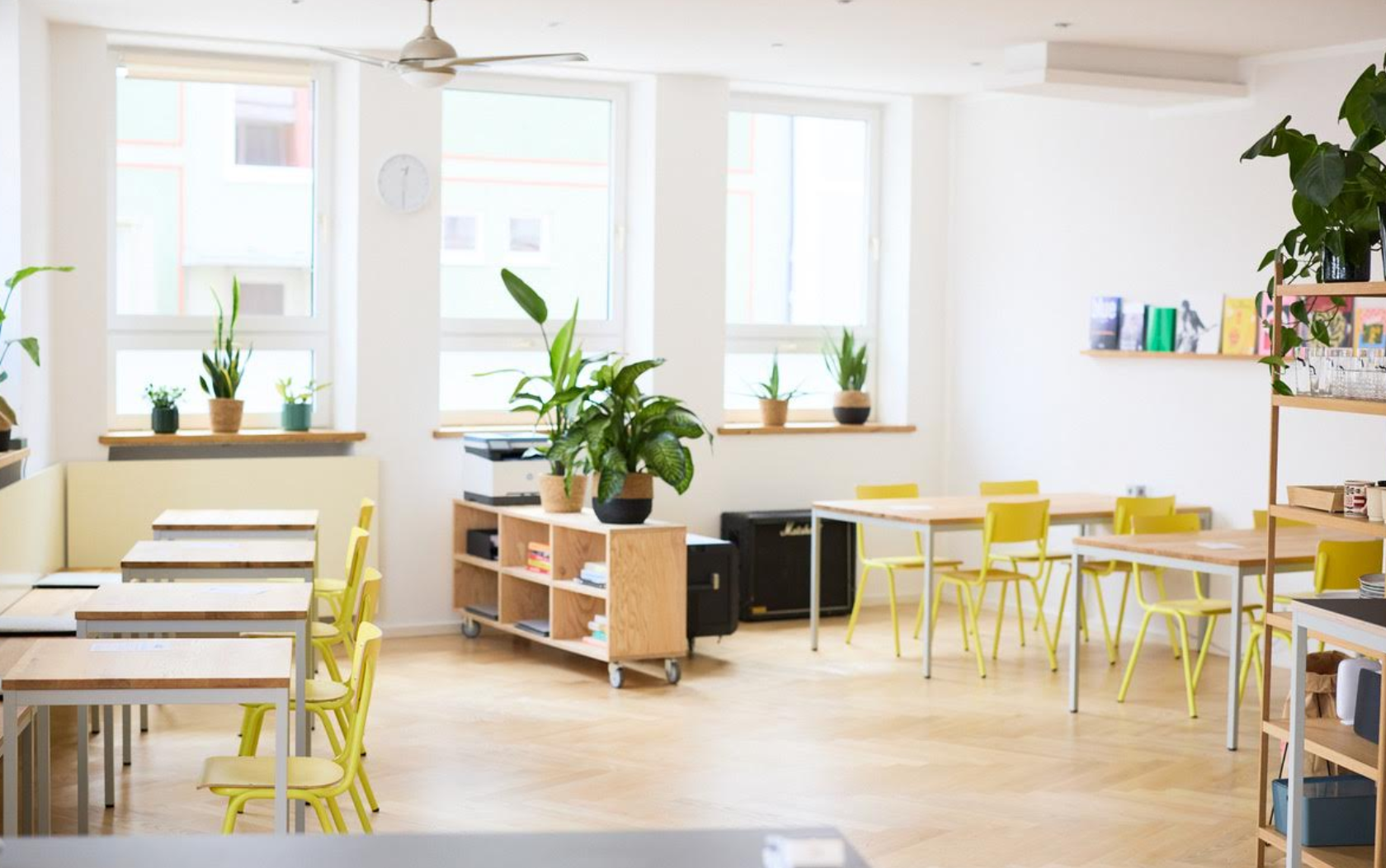 Heller Raum mit großen Fenstern, grünen Pflanzen, gelben Stühlen, Holztischen, Bücherregalen und Sofa. Modern eingerichtet, viel Tageslicht.
