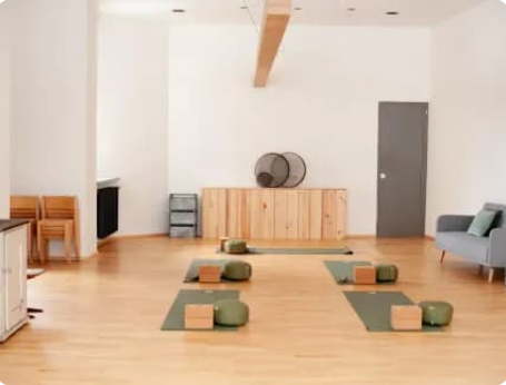 Ein helles, minimalistisch eingerichtetes Zimmer mit Yoga- oder Meditationsmatten, Kissen und Blöcken auf dem Boden, einem kleinen Sideboard gegen die Wand und einem grauen Sessel.