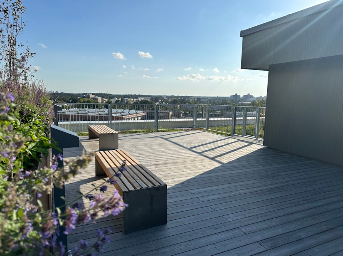 Eine Dachterrasse mit Holzbelag, zwei Sitzbänken, blühenden Pflanzen auf der linken Seite, ein Blick auf eine Stadt im Hintergrund und eine klare, sonnige Himmelsszene.