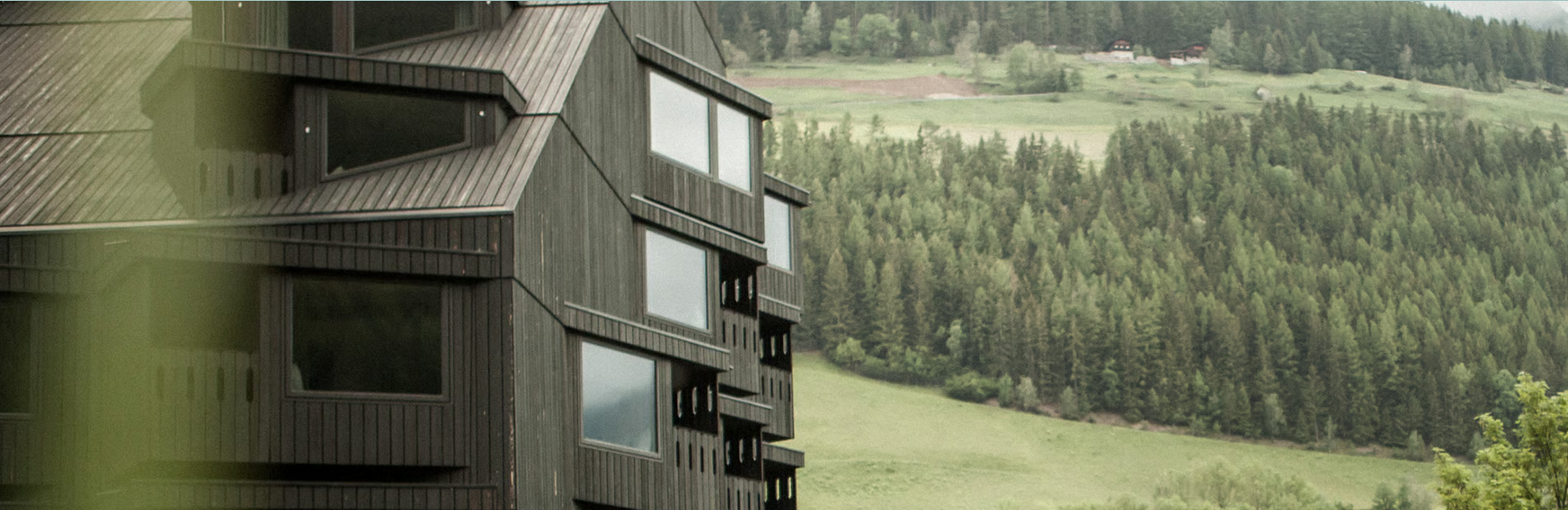 Ein modernes, dunkles Holzhaus in einer ländlichen, grünen Umgebung mit Hügeln und Wäldern im Hintergrund.