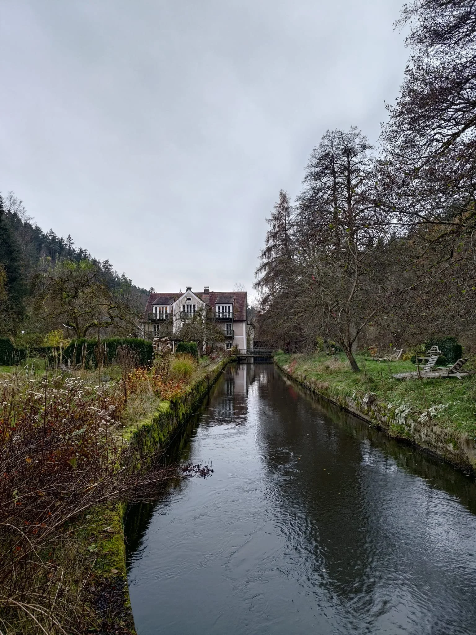 Ein ruhiger Fluss fließt durch eine grüne Landschaft mit Bäumen und einem modernen Haus im Hintergrund, das am Ufer steht.
