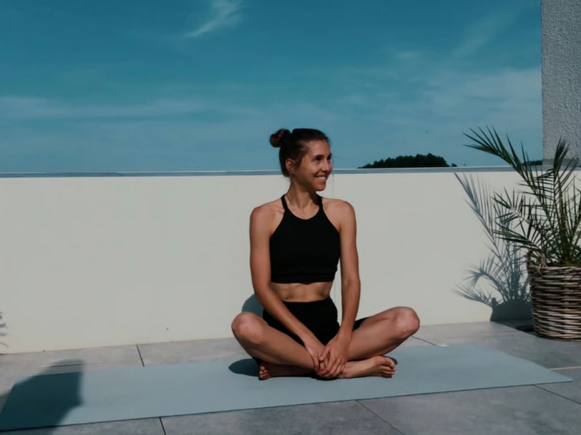 Junge Frau in Schwarz sitzt im Lotussitz auf Yoga-Matte auf einer Terrasse mit Meer im Hintergrund, umgeben von Pflanzen unter blauem Himmel.