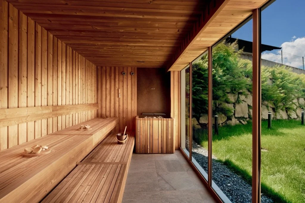 Innenansicht einer Sauna mit Holzverkleidung und Panorama-Windows, Blick ins grüne Freie mit Büschen und Himmel.