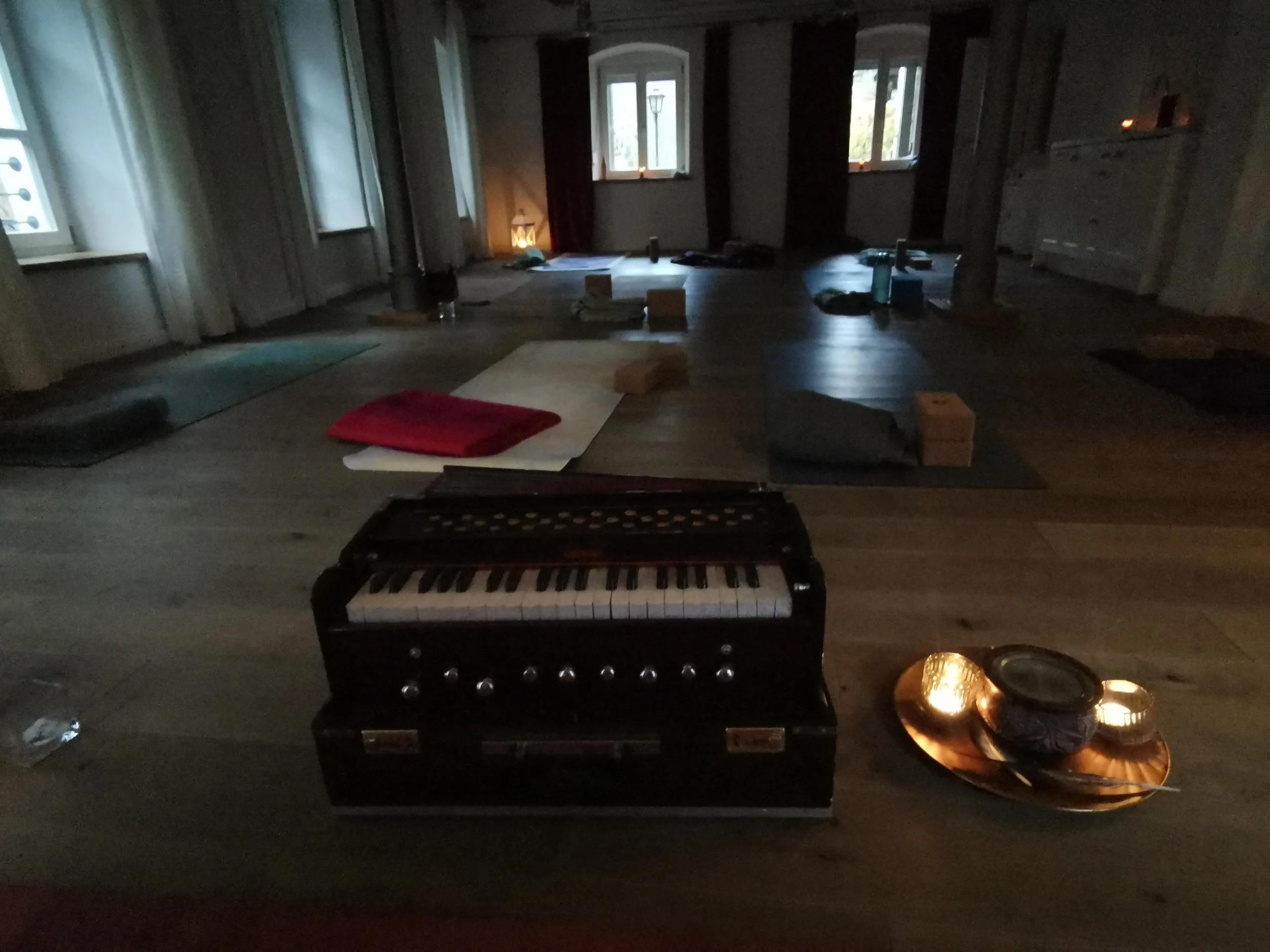 Ein dunkler Raum mit mehreren Fenstern, Kerzen, einem Klavier, Yogamatten, Wasserflaschen und weiteren Gegenständen auf dem Boden, vermutlich für eine Yoga- oder Meditationssitzung vorbereitet.