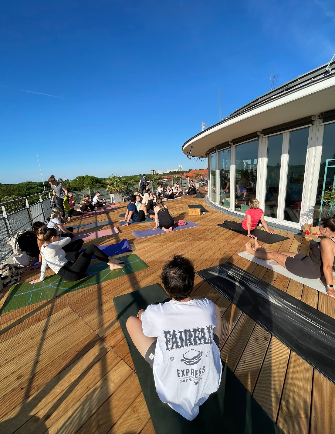 Menschen, die auf einer Dachterrasse Yoga machen bei sonnigem Wetter, mit Blick auf den Himmel und die Stadt im Hintergrund.