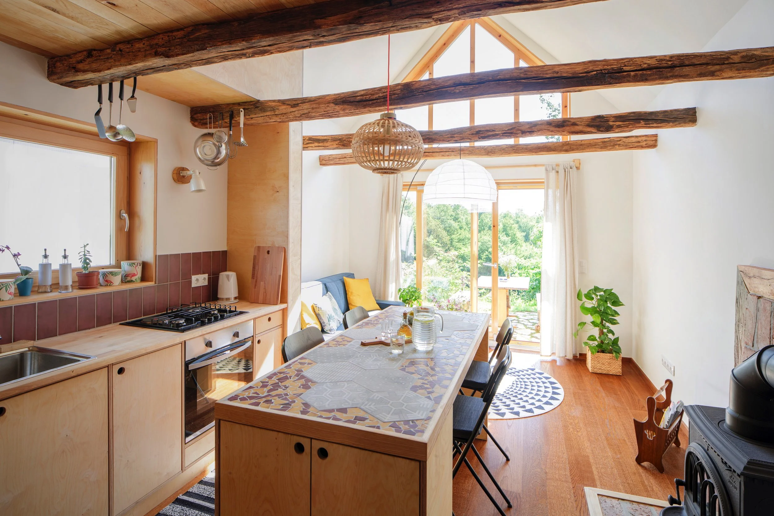 Ein heller, gemütlicher Wohn- und Essbereich mit sichtbaren Holzdeckenbalken, großen Fenstern und einem Esstisch mit Stühlen. Im Hintergrund sind Zimmereinrichtungen und Pflanzen sichtbar.