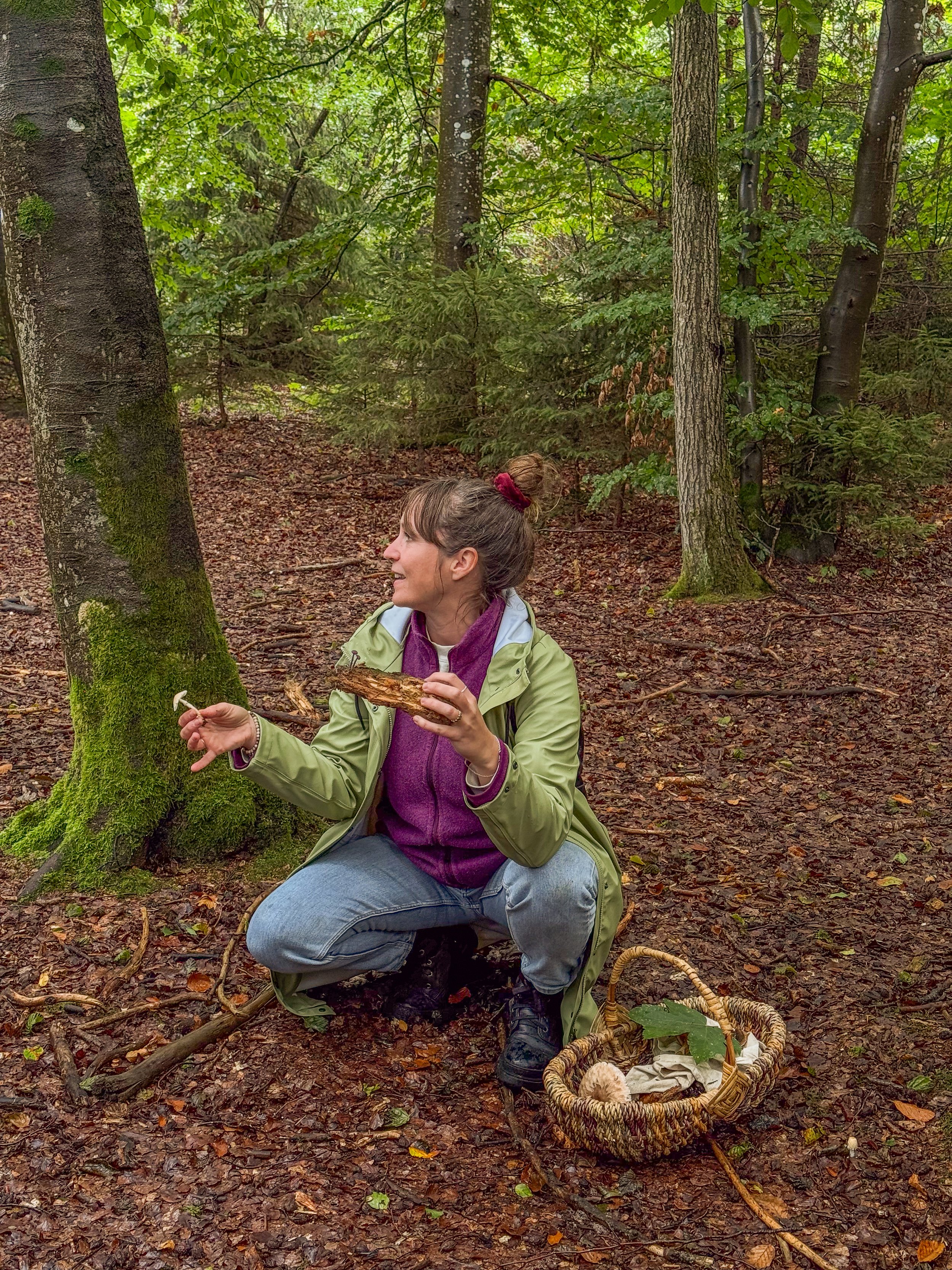 Eine junge Frau sitzt im Wald auf den Knien, hält ein Stück Baumrinde und spricht. Sie trägt eine grüne Jacke, eine lila Fleece, Jeans und feste Schuhe. In der Nähe liegt ein Korb mit Pilzen, um sie zu sammeln.