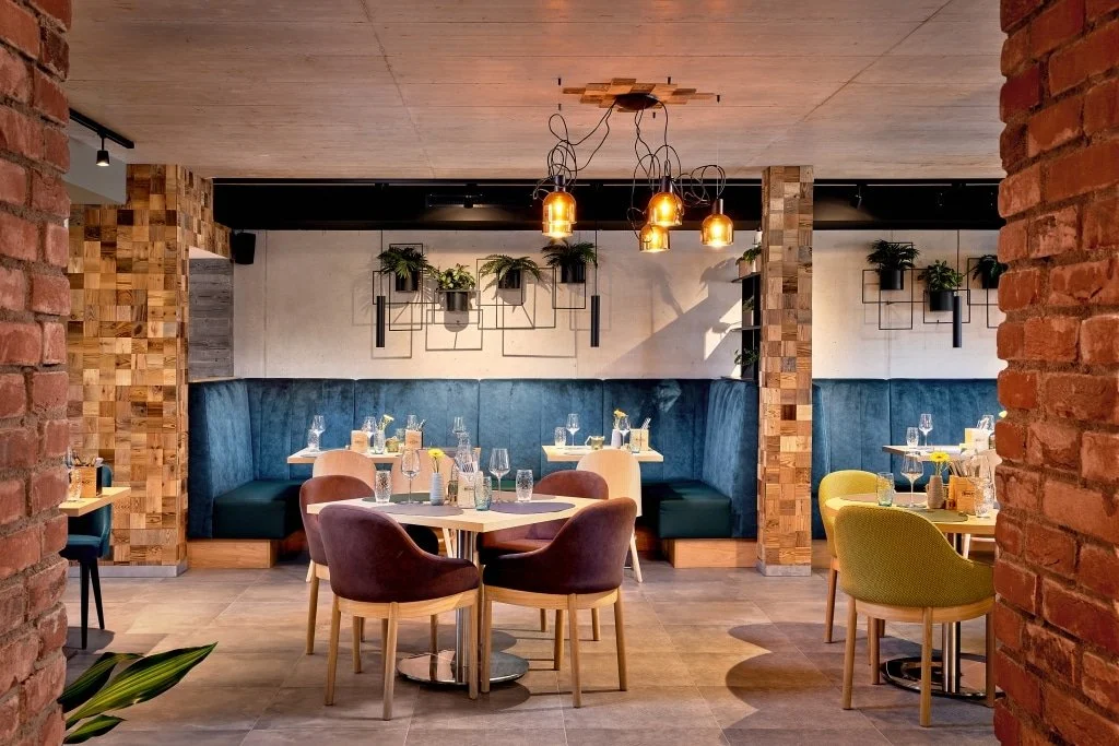Ein modern eingerichteter Restaurantraum mit Holzwanddekoration, blauen und grünen Sitzbanksofas, bunten Stühlen, Tischen mit Gläsern, Wasser und Kerzen sowie einer warmen Beleuchtung.