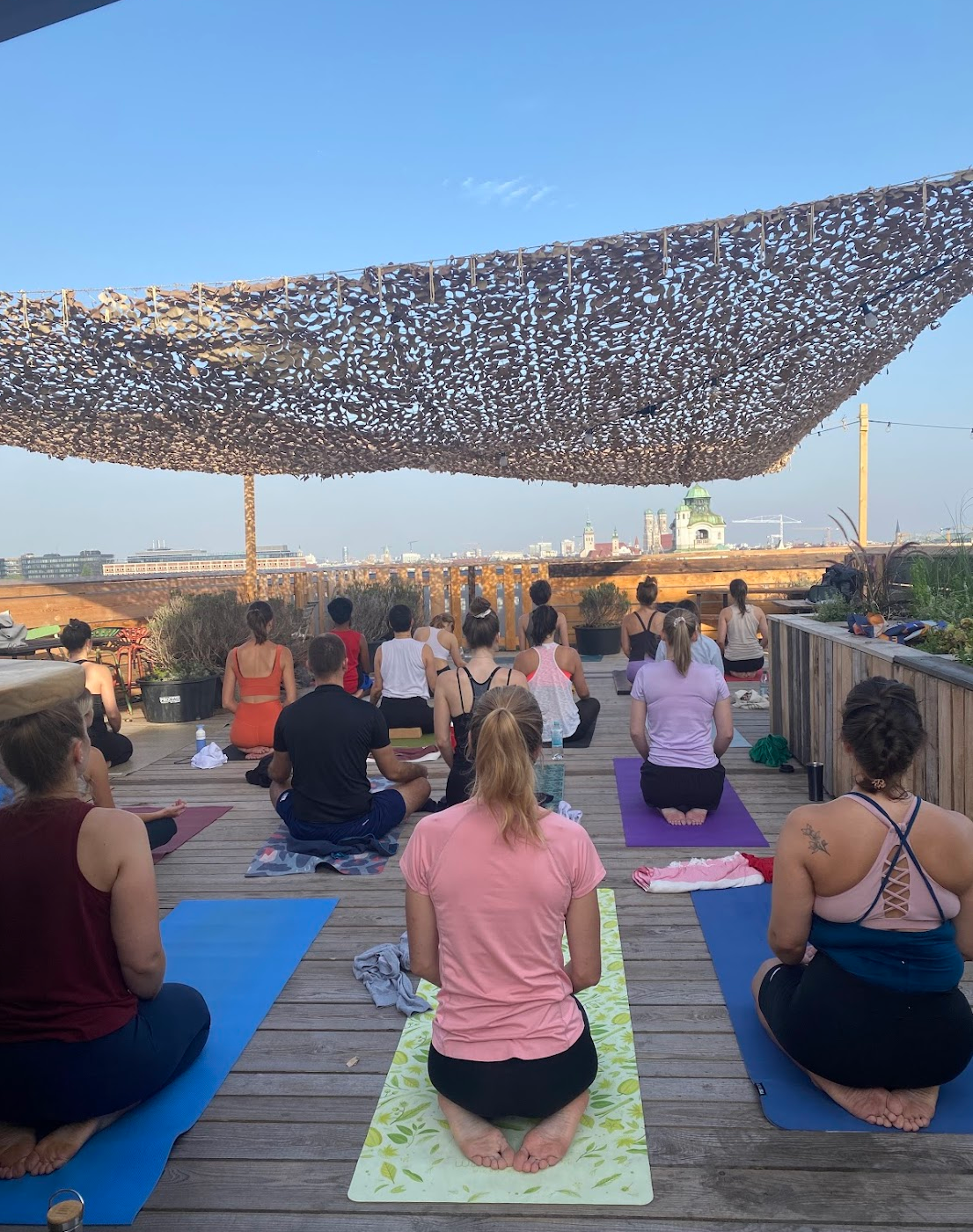 Menschen, die auf einer Dachterrasse im Yoga-Kurs sitzen, mit Blick auf die Stadt, unter einem Netztuch am Himmel.