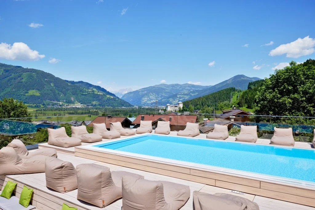 Ein Pool mit bequemen Sitzsäcken auf einer Terrasse mit Blick auf grüne Hügel und Berge unter einem blauen Himmel.