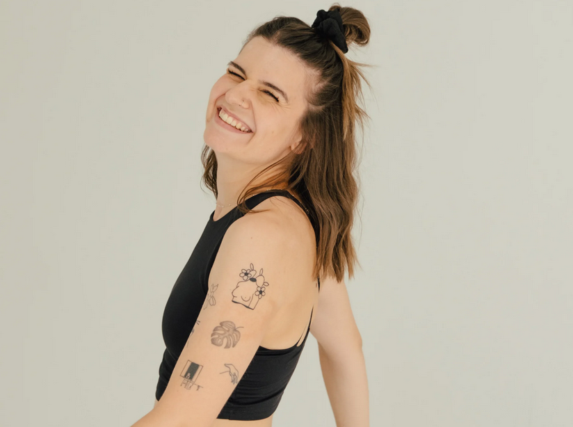 Lächelnde junge Frau mit Tattoos an Arm, trägt schwarzes Top, hairstyle in Zopf, steht vor hellem Hintergrund.