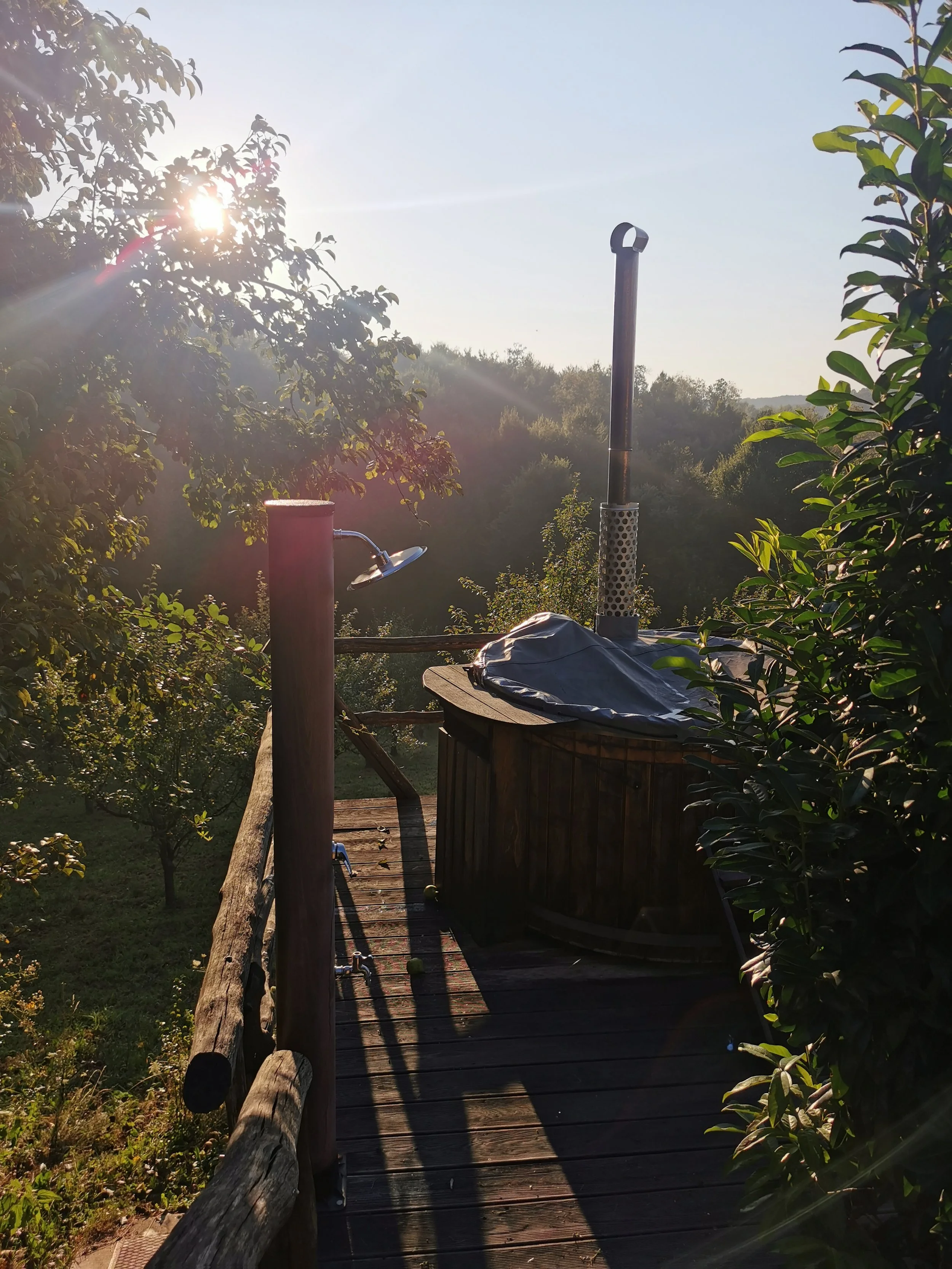 Blick auf einen holzbeplankten Balkon im Freien, umgeben von Bäumen, mit Sonnenschein im Hintergrund. Auf dem Balkon steht ein Heizstrahler und ein Holzkasten, vermutlich ein heißer Wassertank, bedeckt mit einer schwarzen Plane.