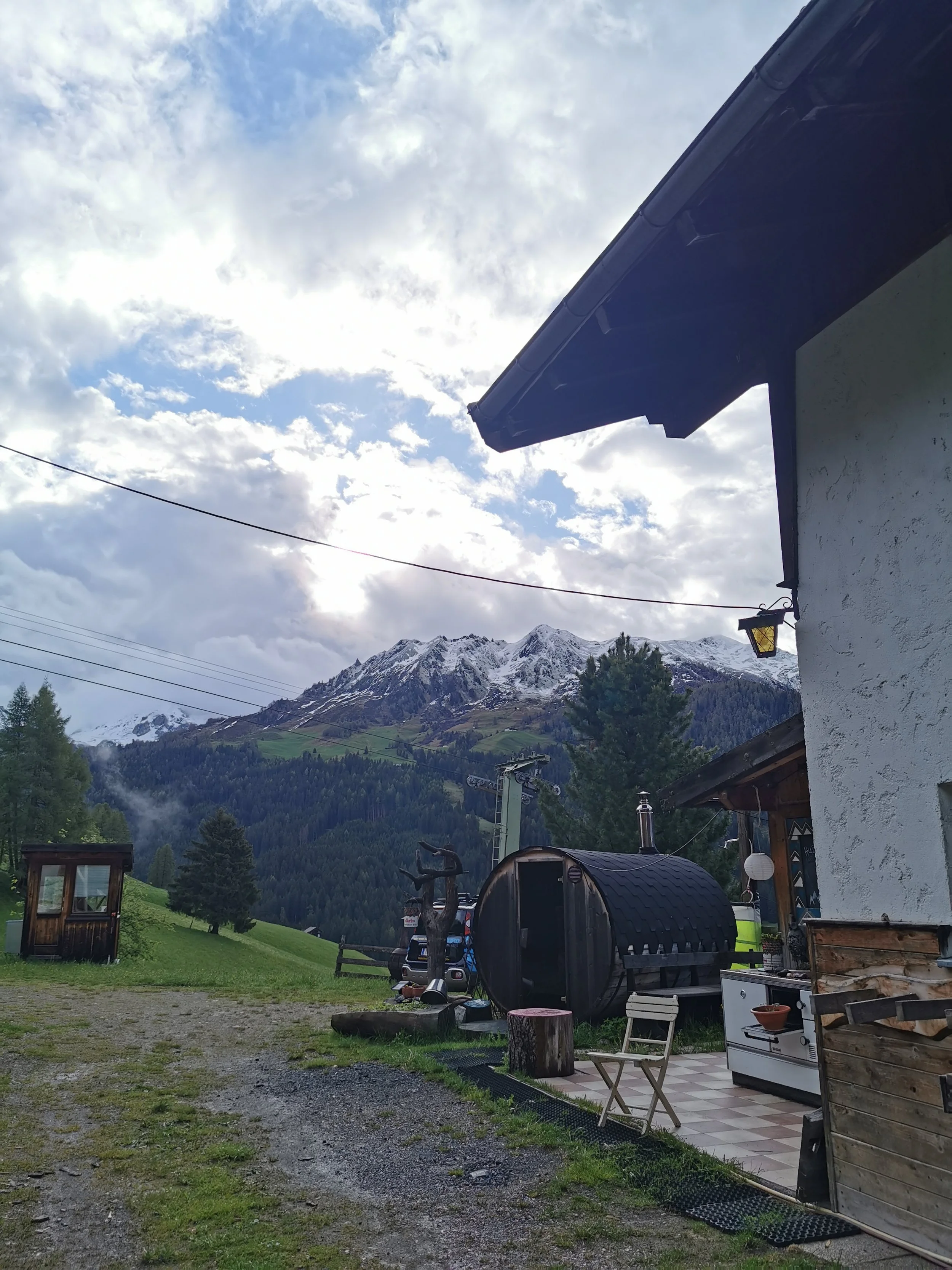 Berglandschaft mit schneebedeckten Gipfeln im Hintergrund, Haus mit Terrasse im Vordergrund, Garten mit Möbeln, Holzofen und Bäumen, bewölkter Himmel.