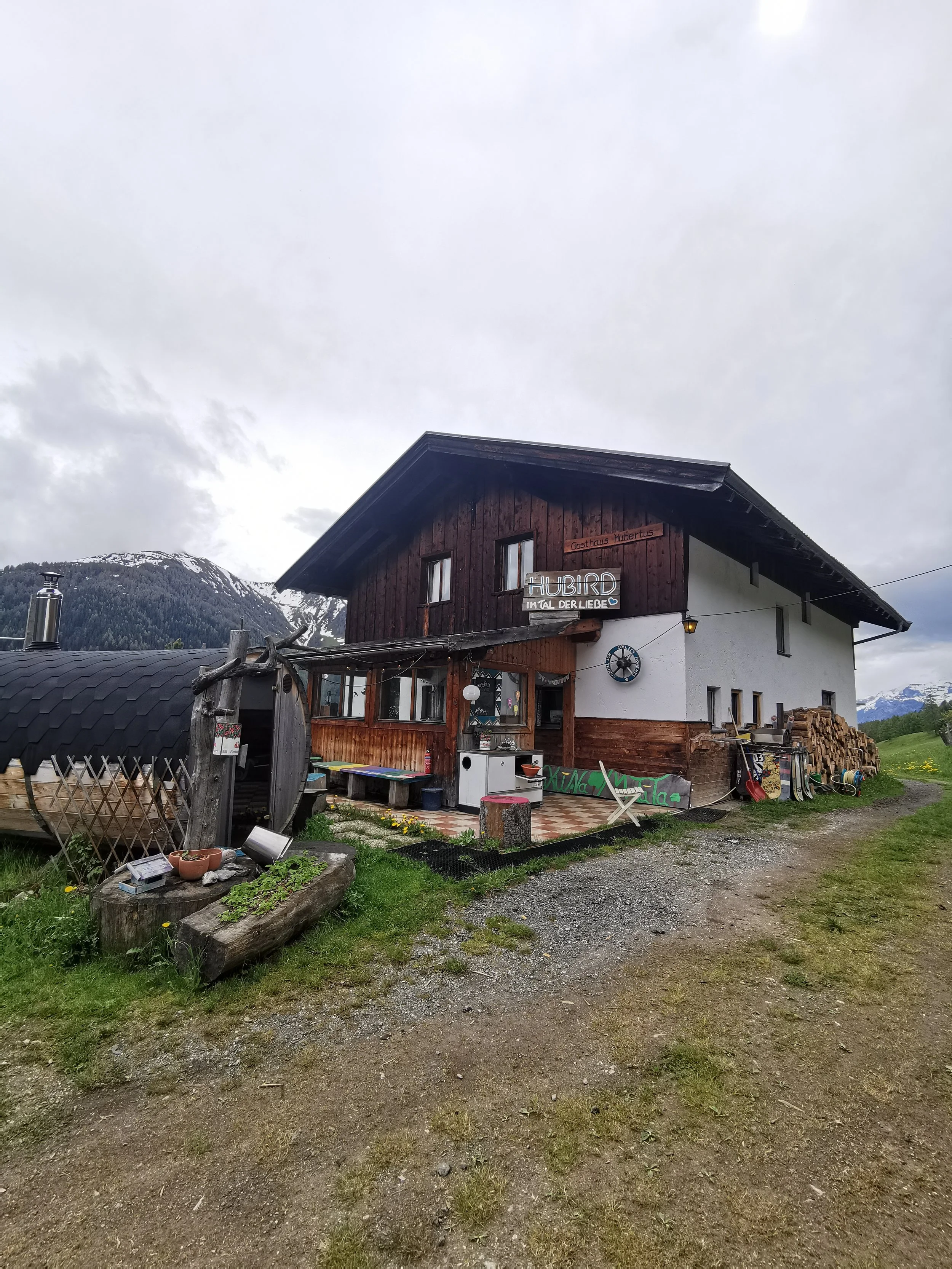 Ein Haus mit einem Holz- und Putz-Fassadenzimmer in einer bergigen Gegend bei bewölktem Himmel, mit draußen liegendem Holz und Gartengeräten.
