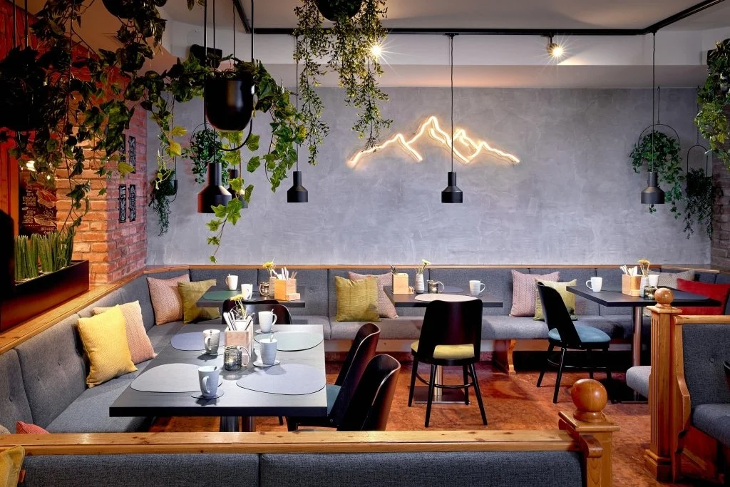 Gemütliches Café mit grauen und bunten Sitzgelegenheiten, grünen Pflanzen, schwarzen Pendelleuchten und einer Neon-Hängekunst am Wand.