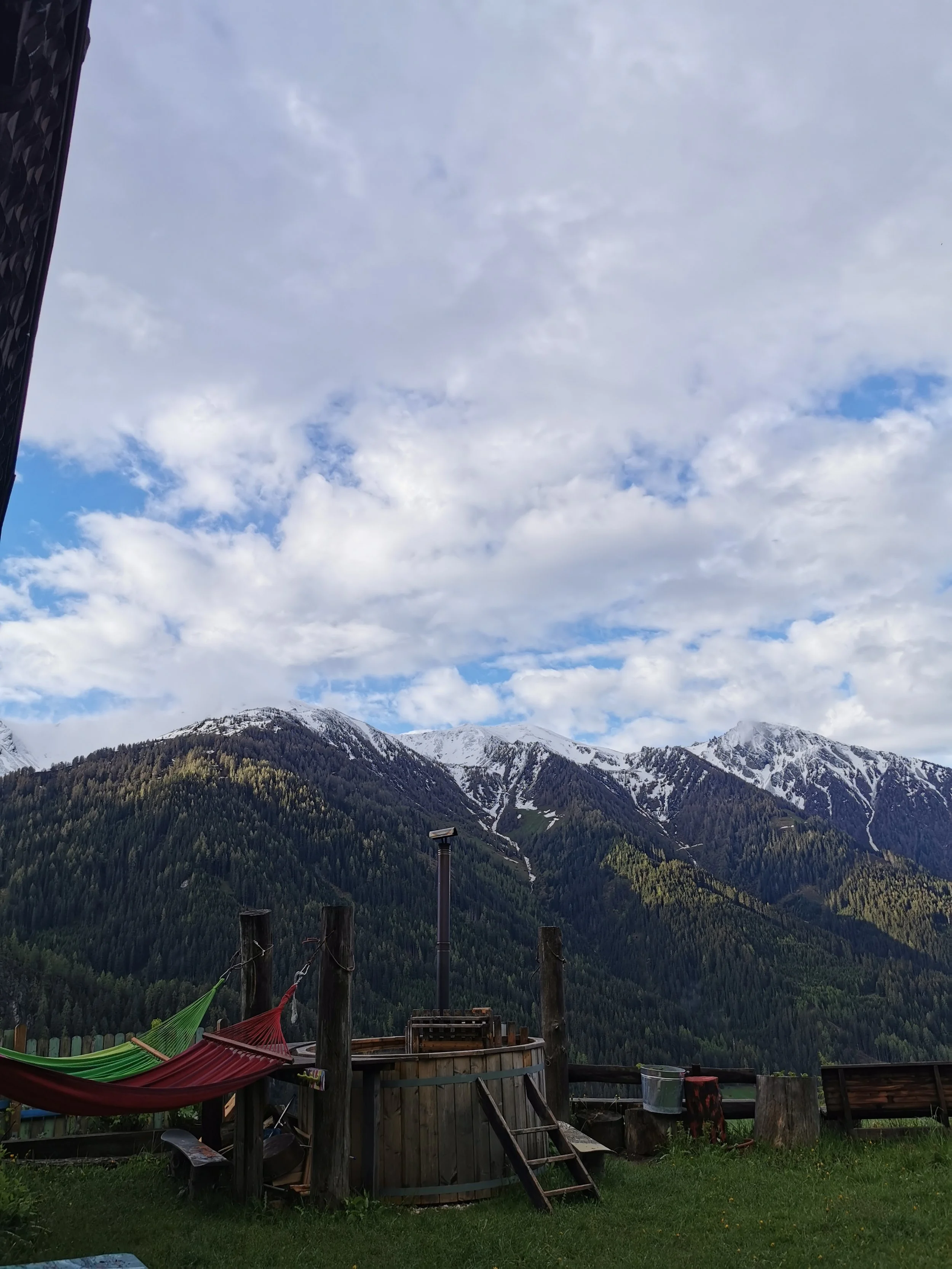 Blick auf eine Bergkulisse mit schneebedeckten Gipfeln, unter einem bewölkten Himmel. Im Vordergrund befindet sich eine Terrasse mit bunten Hängematten, Holzpfosten, und Gartenmöbeln.
