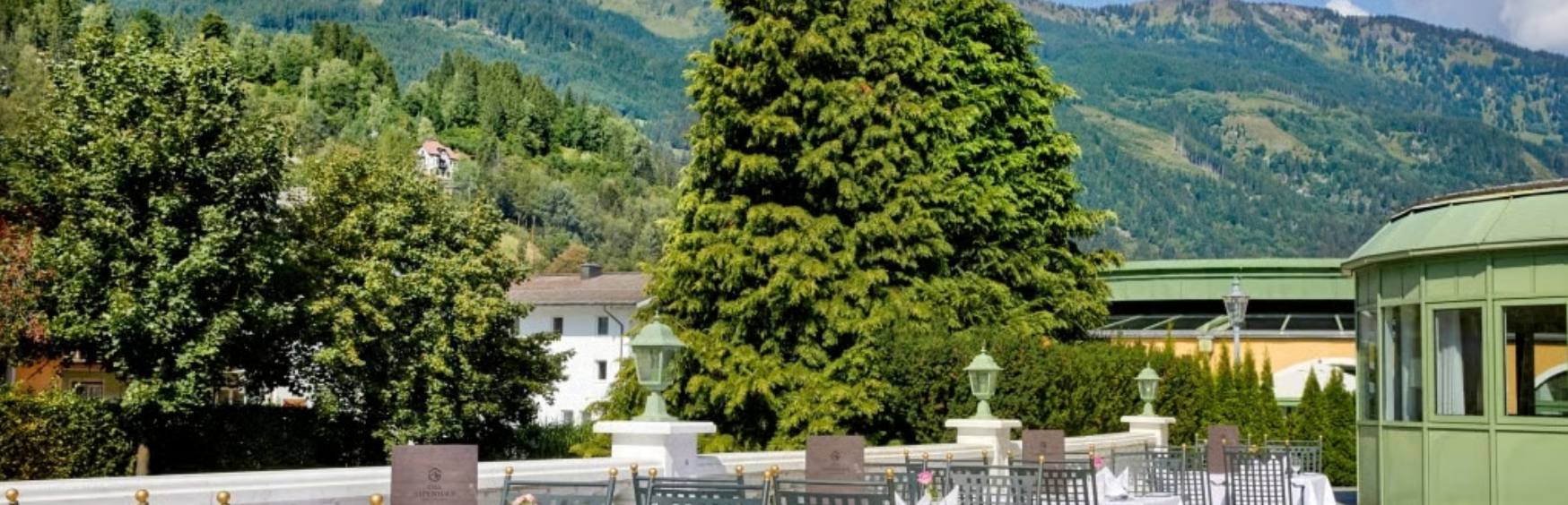 Garten mit großen Bäumen, ziegelgedeckte Dächer im Hintergrund, grüner Pavillon, große Bäume, Laternen am Geländer, Tische und Stühle im Freien.