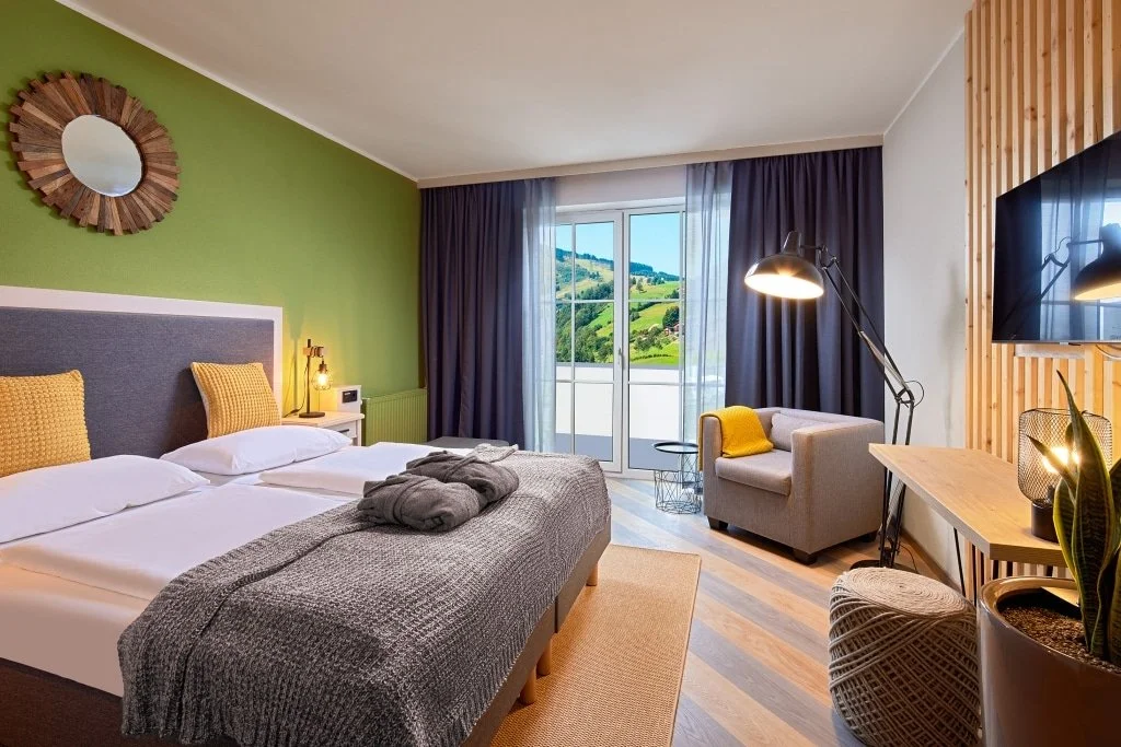 Ein modern eingerichtetes Hotelzimmer mit Doppelbett vor einer grünen Wand, Nachttischen, Stehlampe, Sessel mit gelbem Kissen, Balkon mit Blick auf grüne Hügel, Fernseher an einer Holzverkleidung, Tisch mit Pflanze.