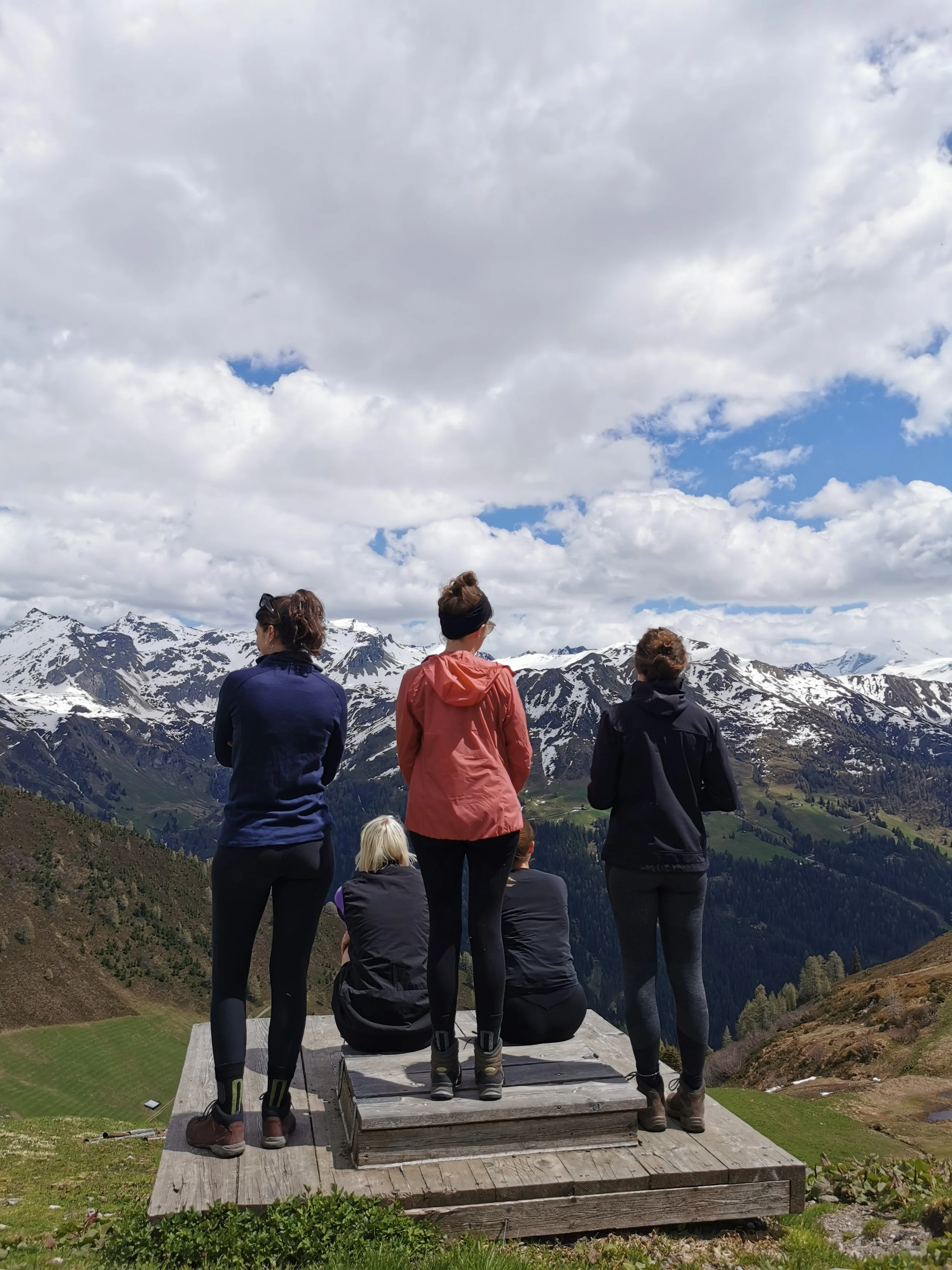Menschen, die auf einer Holzplattform in den Bergen sitzen und stehen, während sie die Aussicht auf schneebedeckte Berge und Wolken im Himmel genießen.