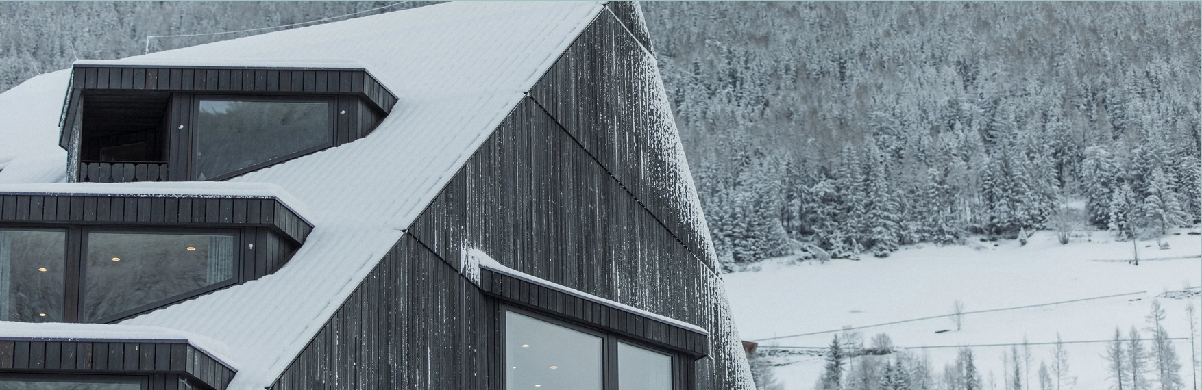 Ein modernes schwarzes Haus mit Schneebedecktem Dach vor einem verschneiten Berg- und Waldhintergrund.