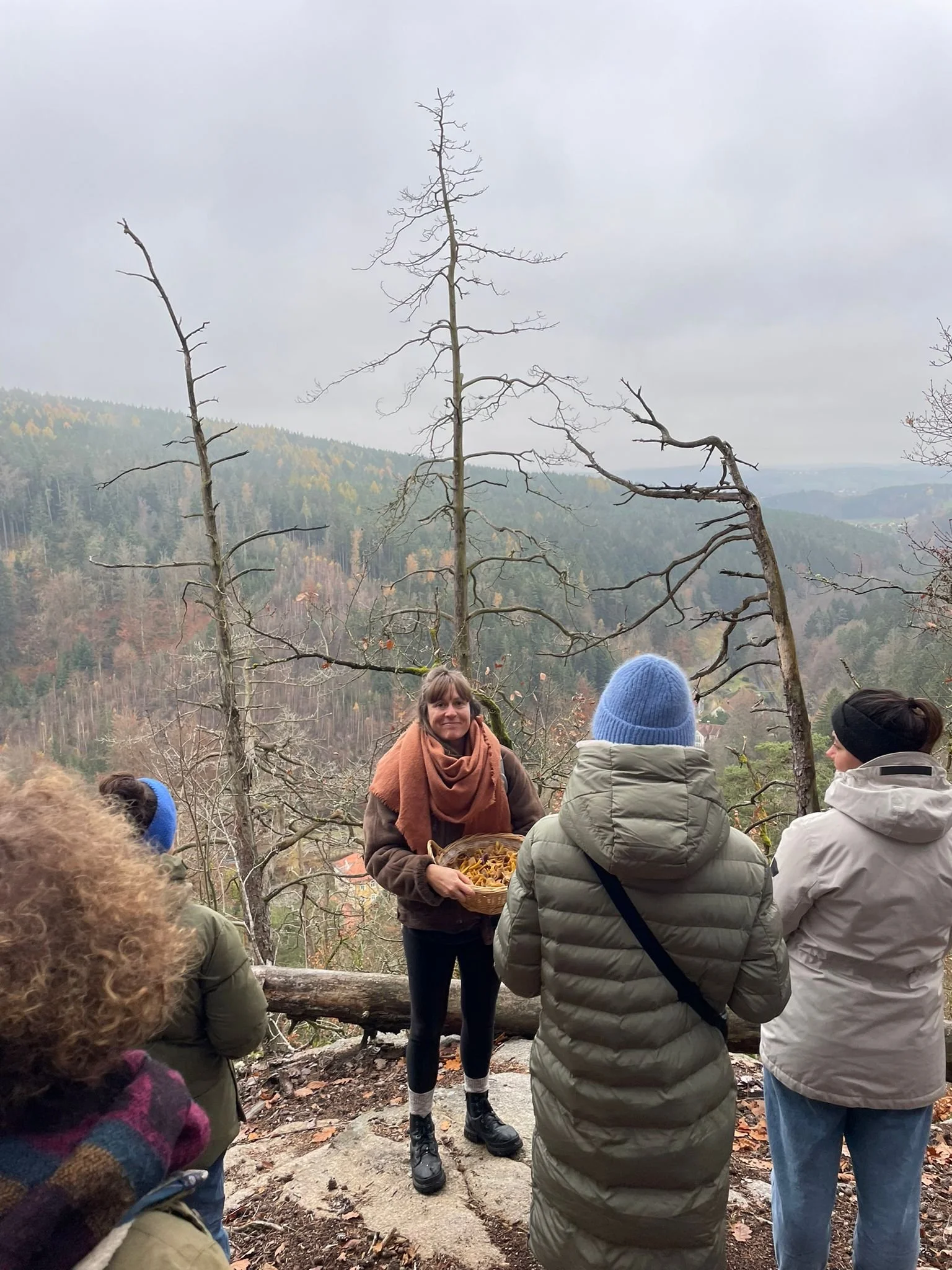 Eine Gruppe von Menschen, die im Freien in einem Wald auf einem Wanderweg stehen. Im Hintergrund sind Bäume und eine nebelige, bewölkte Landschaft zu sehen.