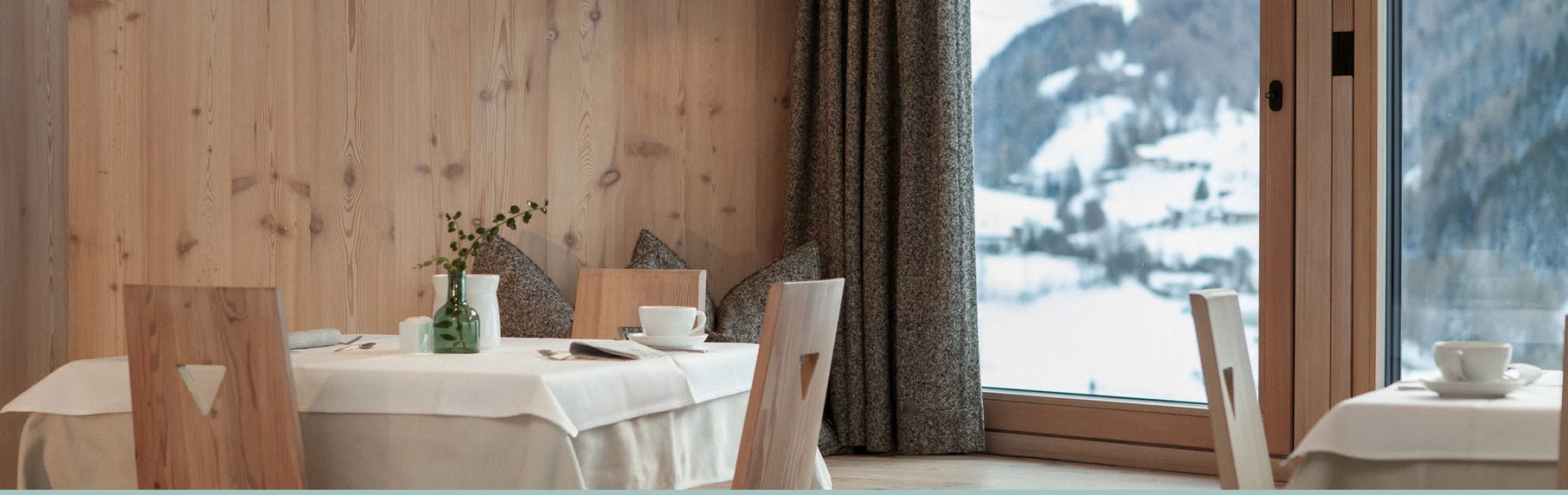 Innenaufnahme eines Restaurants mit Holzwänden, weißen Tischtüchern, weißen Geschirr und einer großen Fensterfront mit Schneelandschaft im Hintergrund.