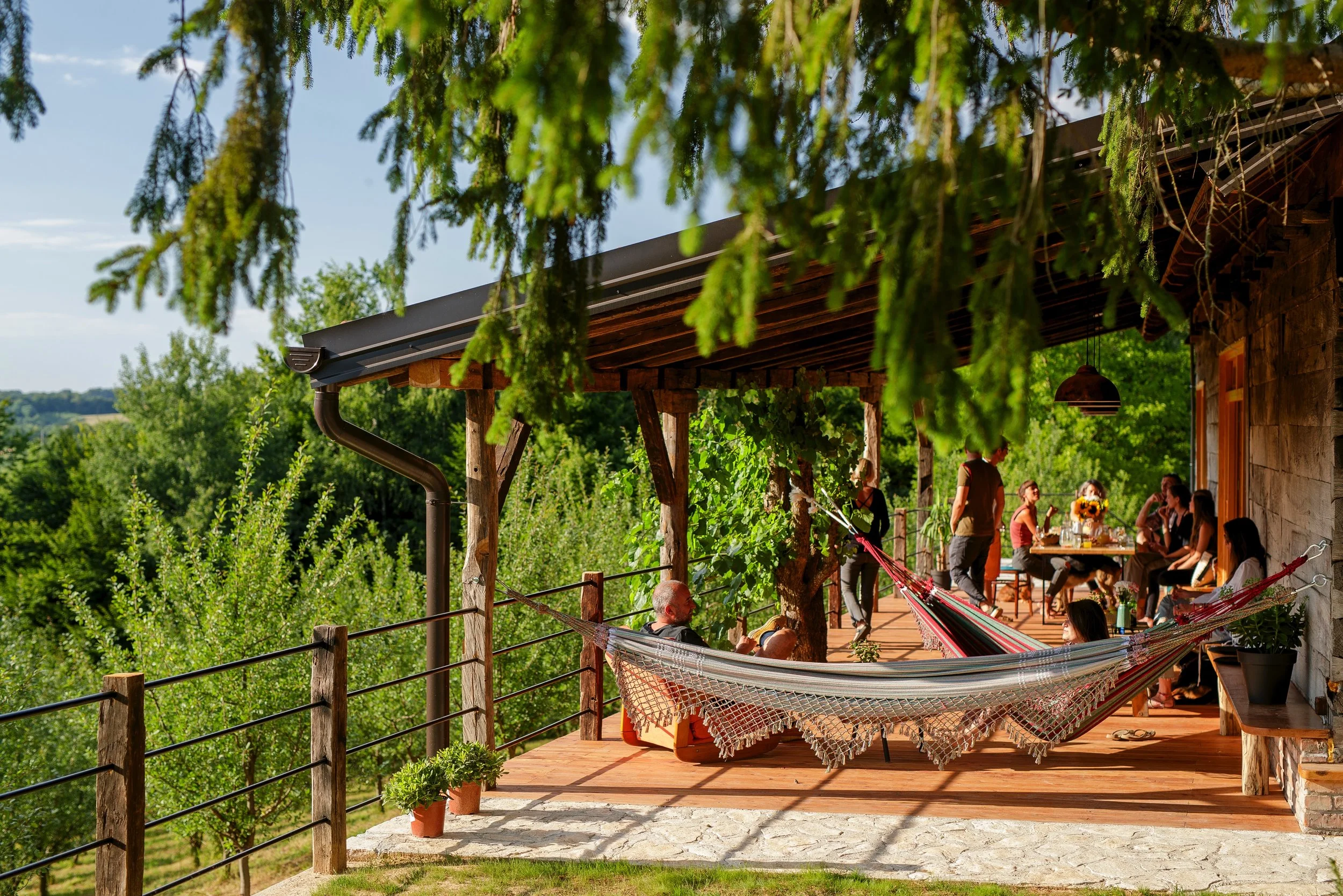 Menschen sitzen und stehen auf einer Holzterrasse vor einem Haus mit Steinfassade, umgeben von grünen Bäumen, bei schönem Wetter. Es gibt Hängematten und eine große Baumkrone im Vordergrund.