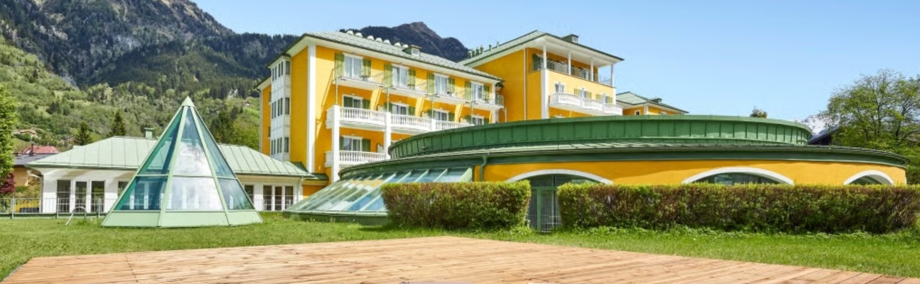 Buntes Hotel mit gelber Fassade, grünen Dächern und zahlreichen Balkonen, umgeben von Bergen und Grünflächen, im Hintergrund ein klarer blauer Himmel.