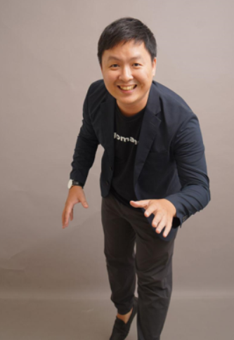 A smiling young man in a black blazer and dark pants posing playfully in front of a plain background.