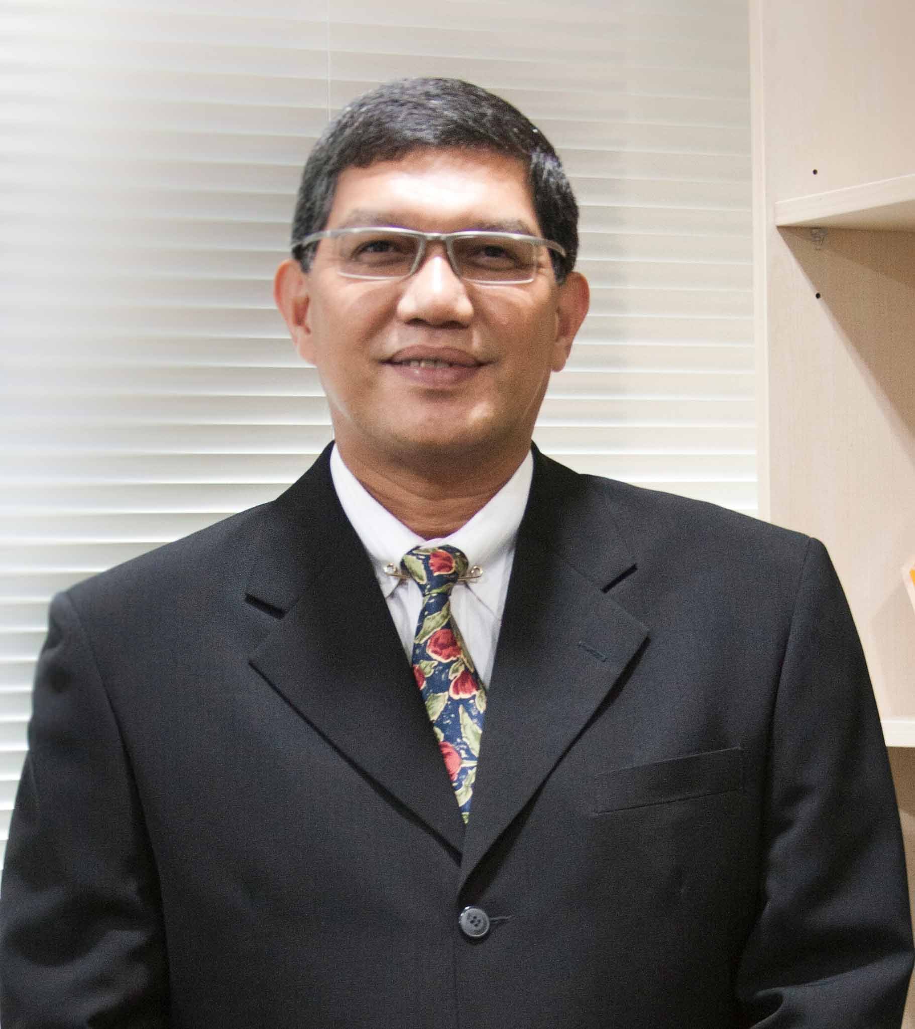 A man wearing glasses, a suit, and a colorful tie, smiling in an indoor setting with blinds and a shelving unit in the background.