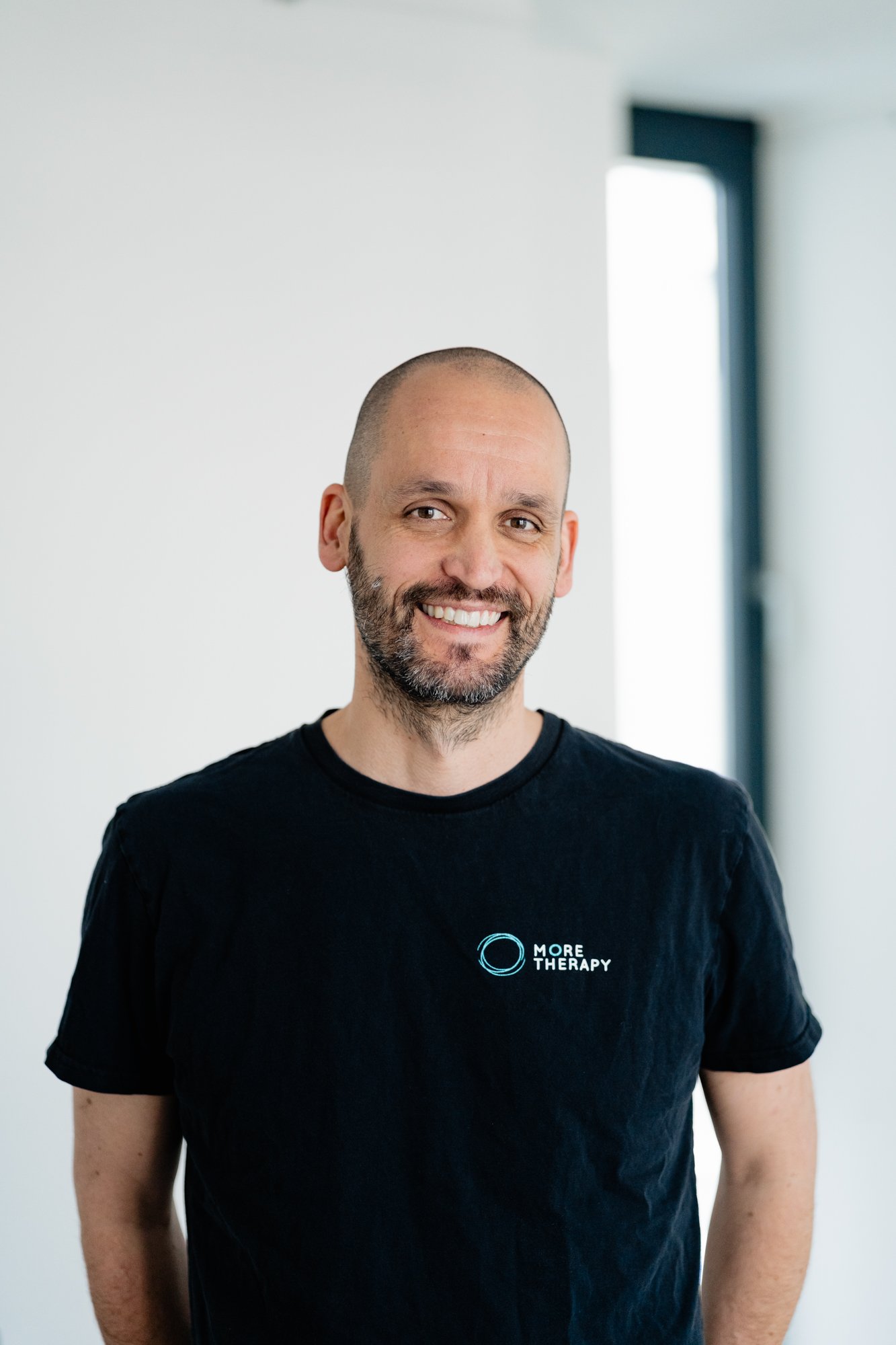 Mann mit kurzen Haaren und Bart, lachend, trägt ein schwarzes T-Shirt mit dem Logo 'More Therapy'.