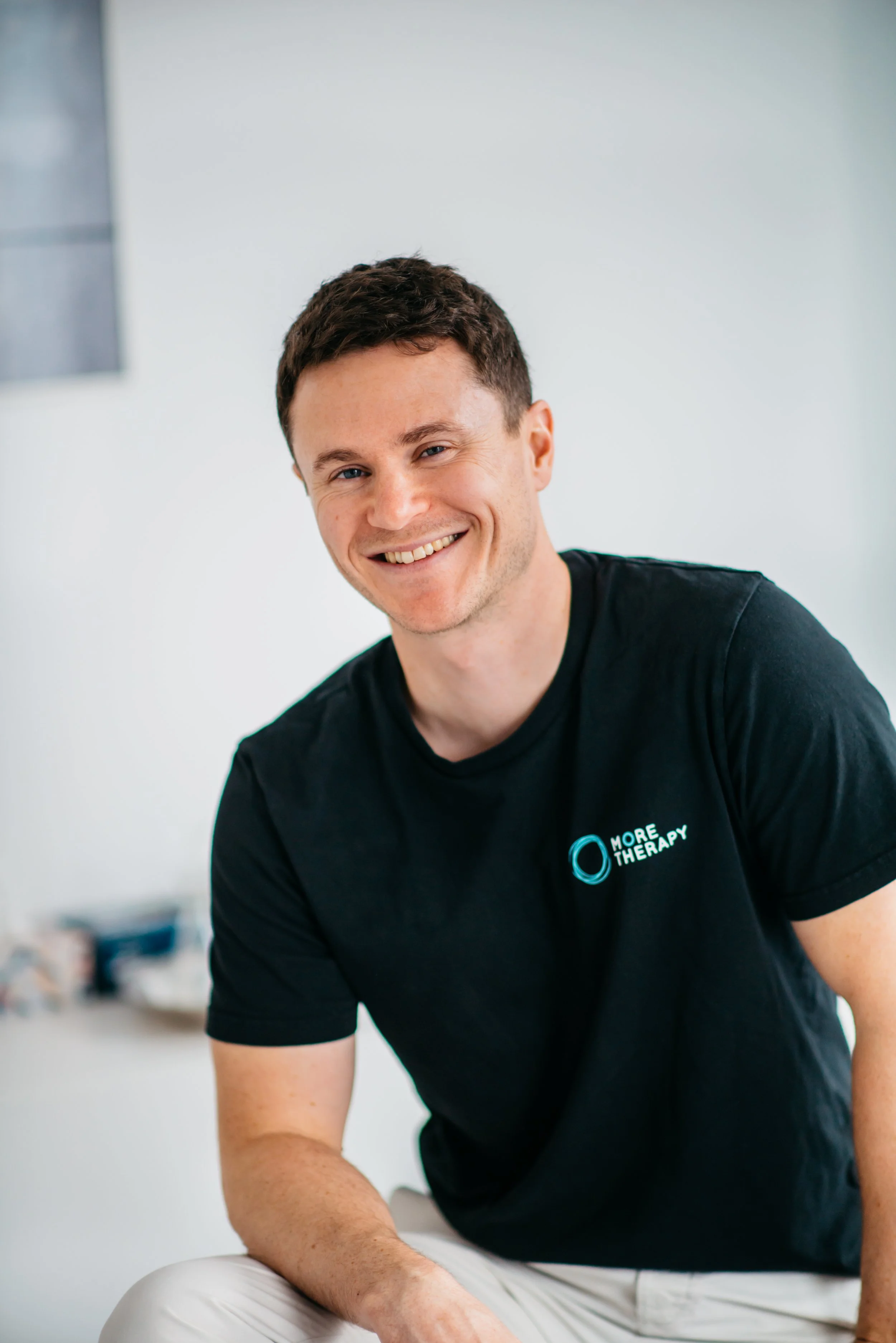 Ein junger Mann lächelt freundlich in die Kamera, trägt ein schwarzes T-Shirt mit dem Logo 'More Therapy' auf der Brust, sitzt in einem hellen Raum.