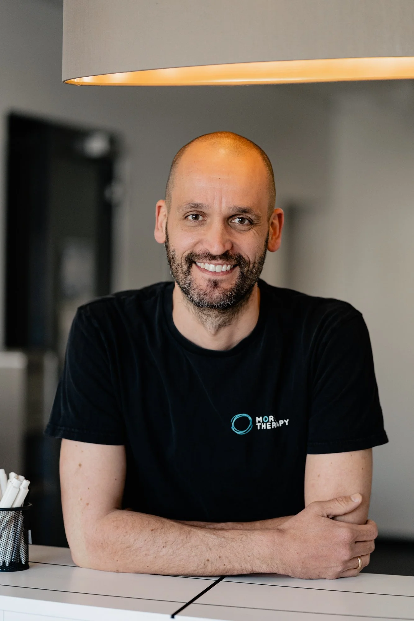 Mann mit kurzen Haaren und Bart, lächelnd, sitzt an einem Tisch im Raum, trägt ein schwarzes T-Shirt mit Logo 'MOR THERAPY'. Osteopath Stuttgart