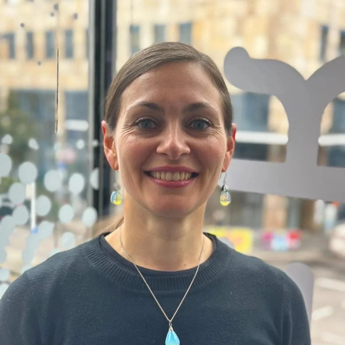 Nahaufnahme einer lächelnden Frau mit braunen Haaren, blauen Augen, Ohrringen und Halskette, im Hintergrund eine Stadt.