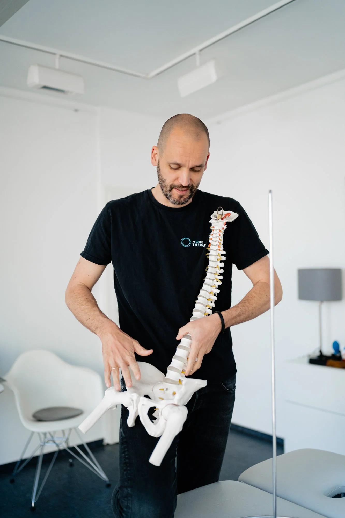 Ein Mann hält ein Modell der menschlichen Wirbelsäule in einem Raum mit weißen Wänden und Stühlen. Malte Wagner. Osteopathie Stuttgart
