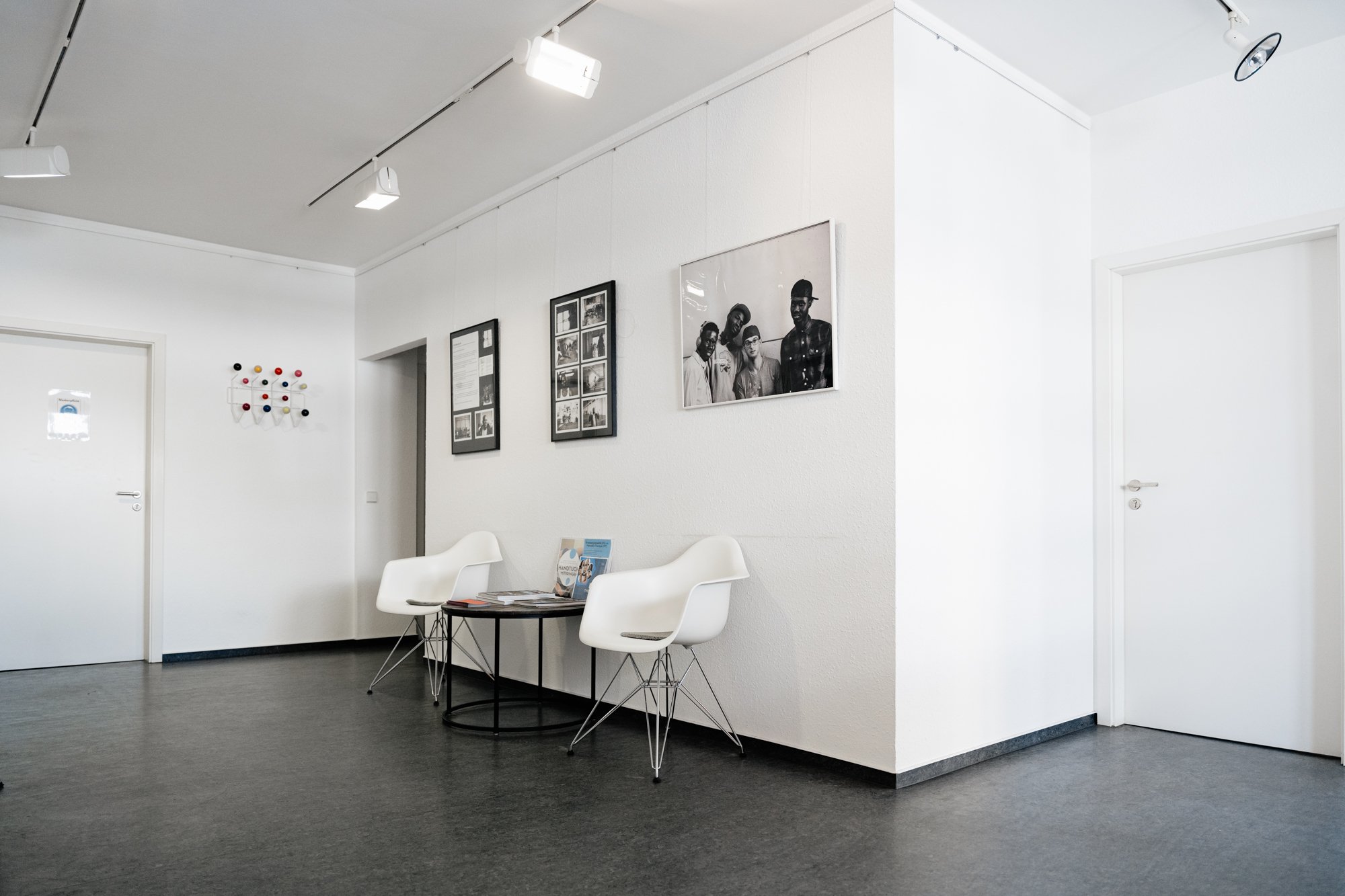Wartezimmer mit zwei weißen Stühlen, einem kleinen schwarzen Tisch mit Zeitschriften, Wandbildern und einer Wanddekoration in einem modernen, minimalistischen Raum. MORE Therapy